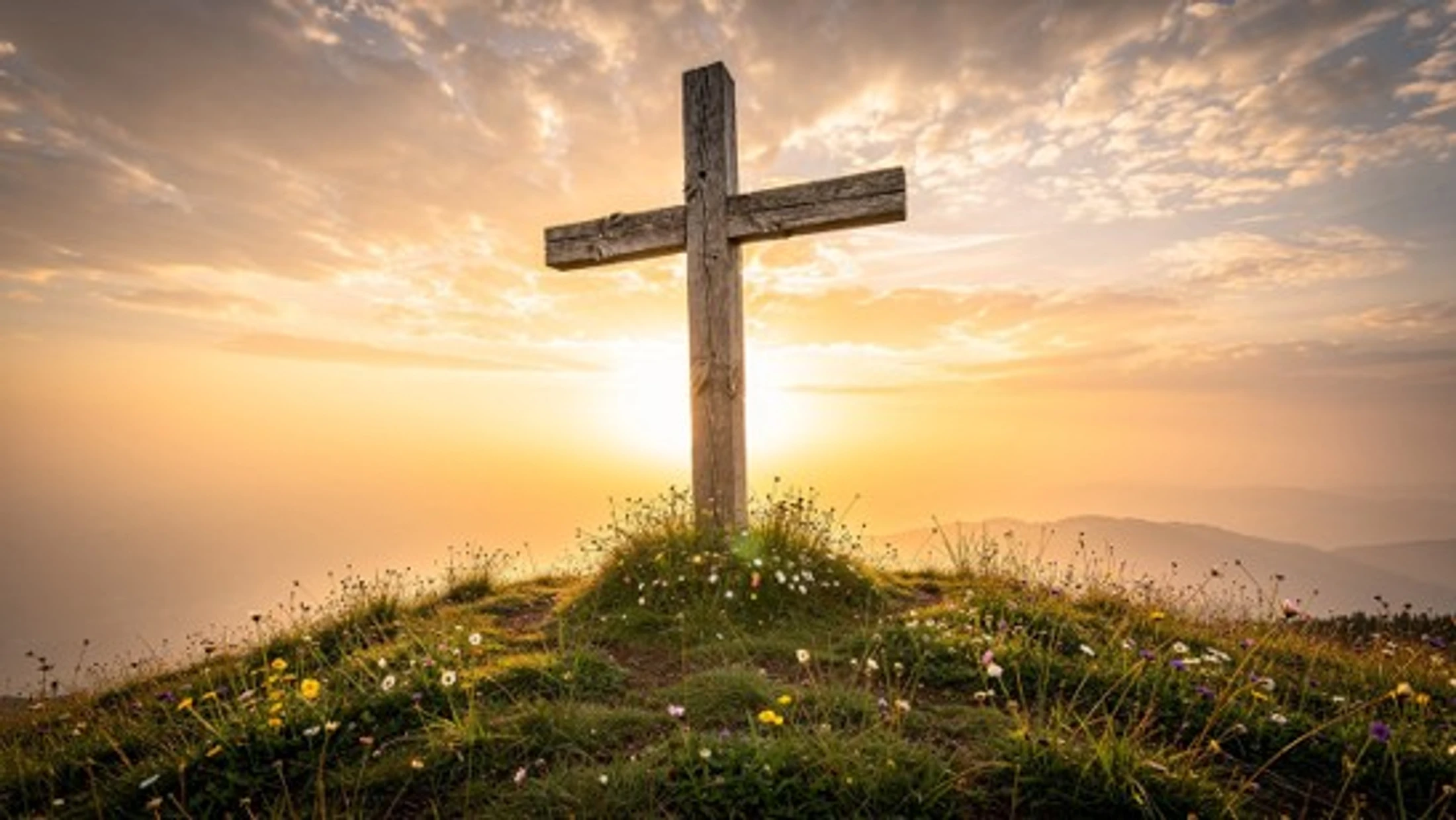 A rustic wooden cross stands on a grassy hilltop with wildflowers, silhouetted against a beautiful golden sunrise with distant mountains in the background.