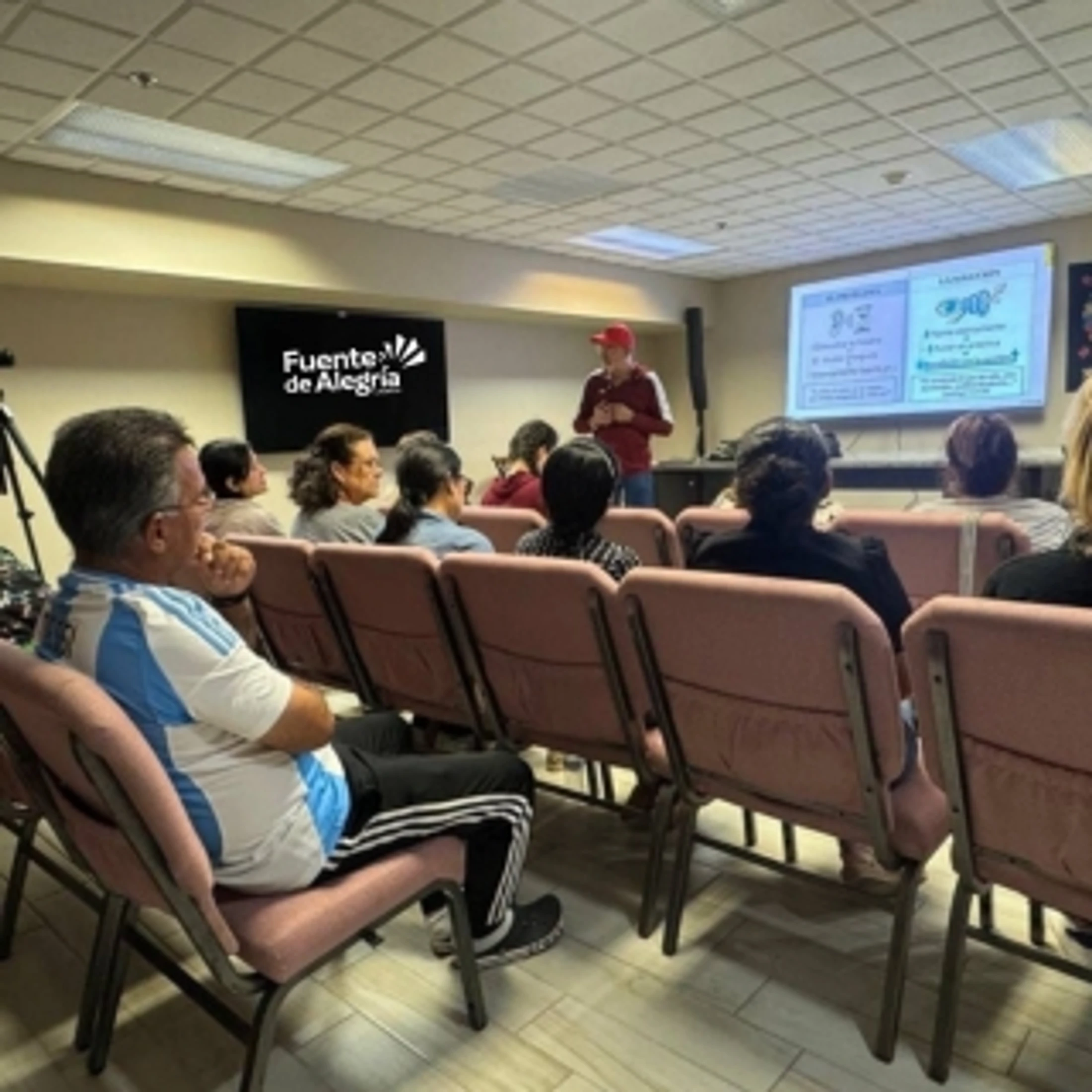 An audience sits in rows of chairs listening to a speaker giving a presentation with a slideshow in a conference room.