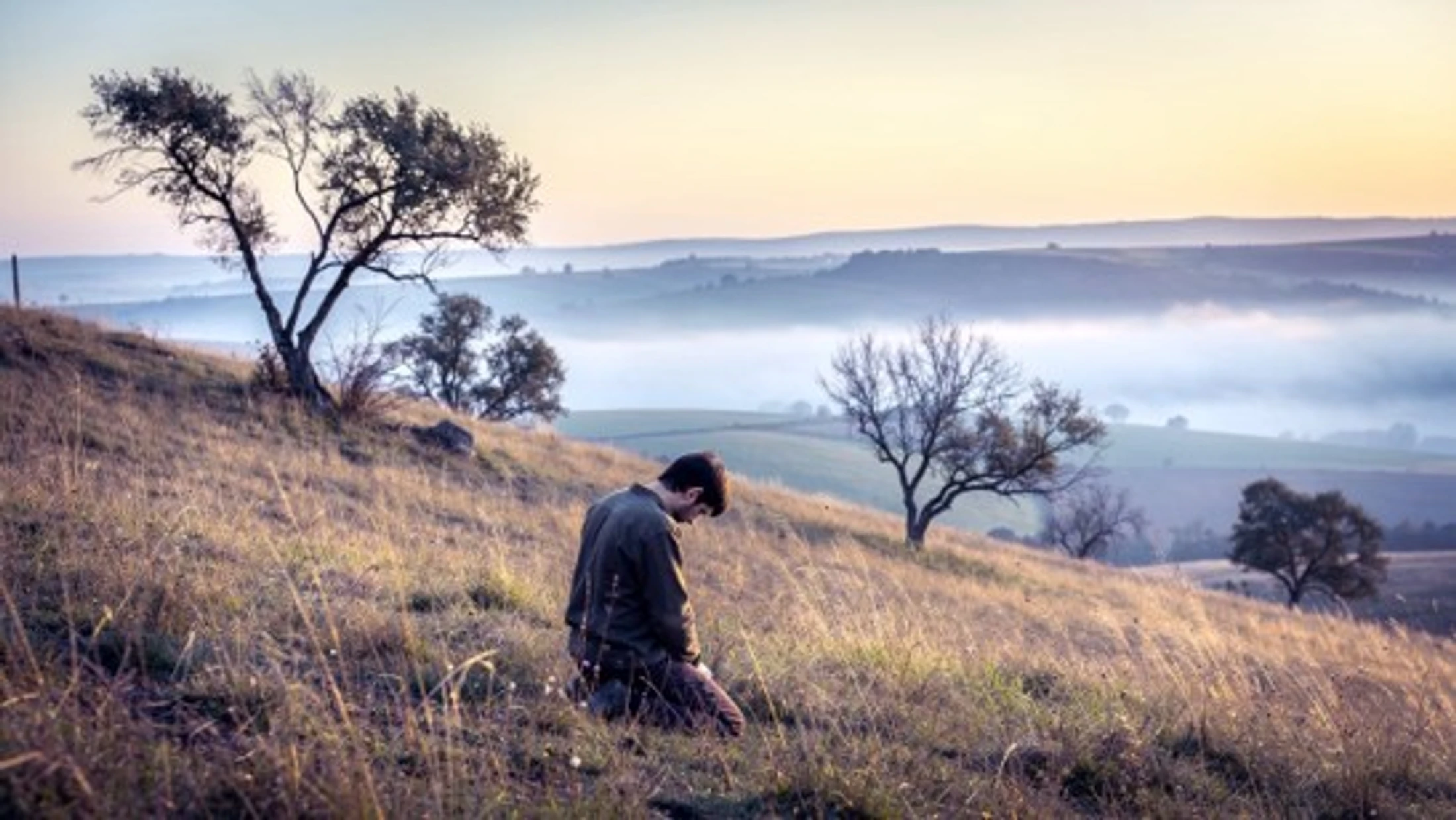 A man kneels with his head bowed on a grassy hillside during a misty sunrise. In the background, rolling hills fade into the fog under a soft sky.