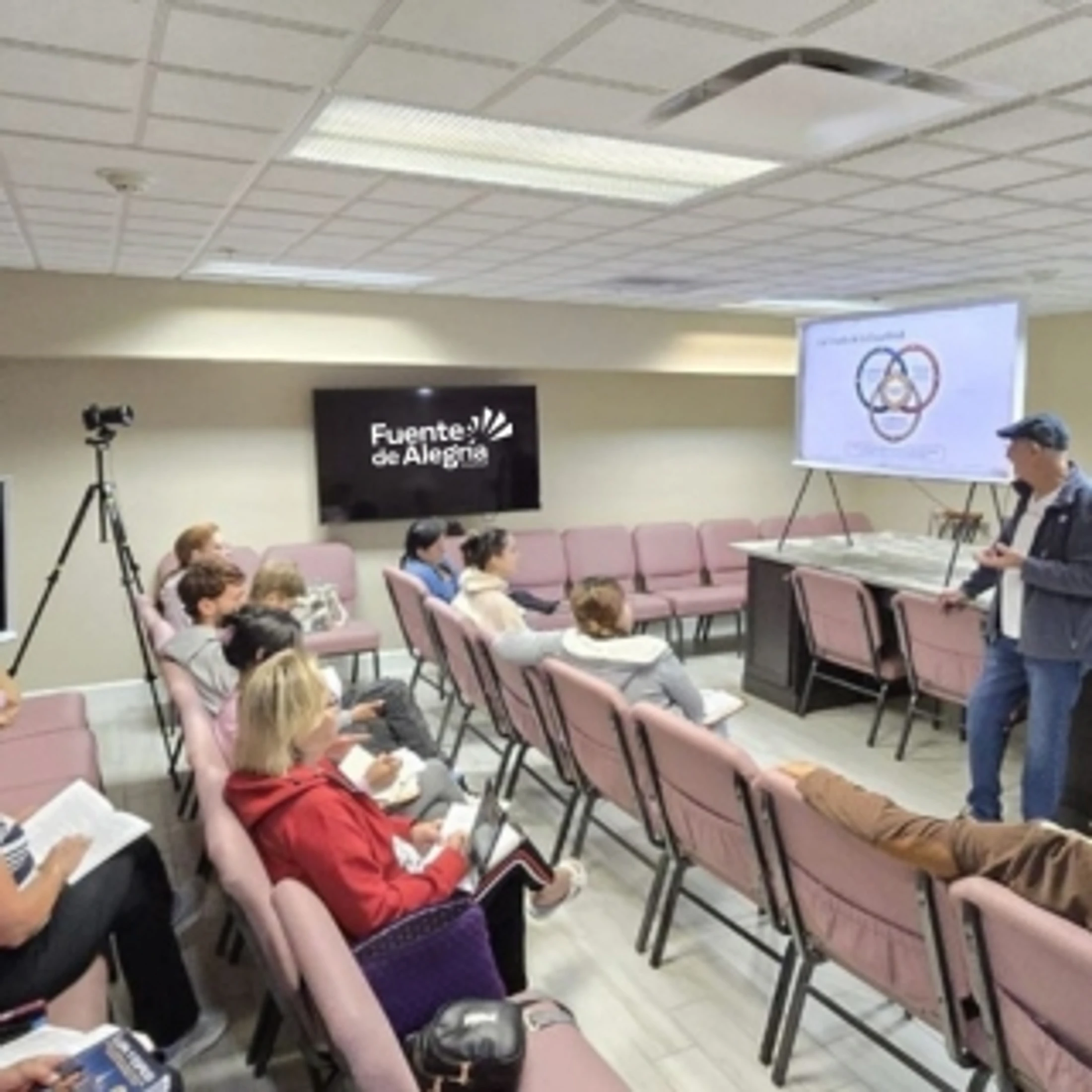 A man in a cap presents a diagram on a whiteboard to a small audience seated in pink chairs in a classroom setting, with a logo on a screen behind them.
