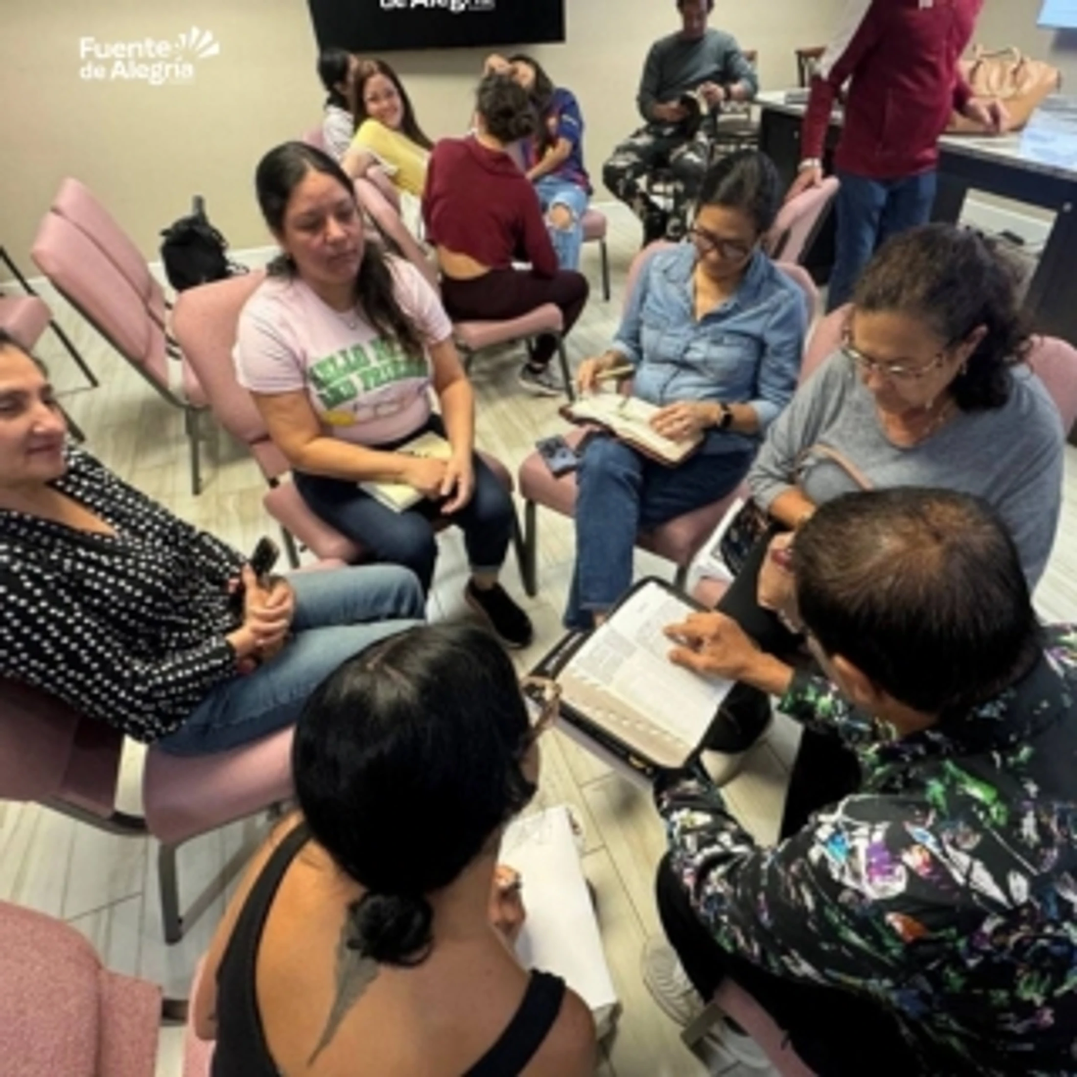 A group of adults sit in a circle on pink chairs, engaged in a discussion while looking at open books, likely Bibles, during a study group meeting.