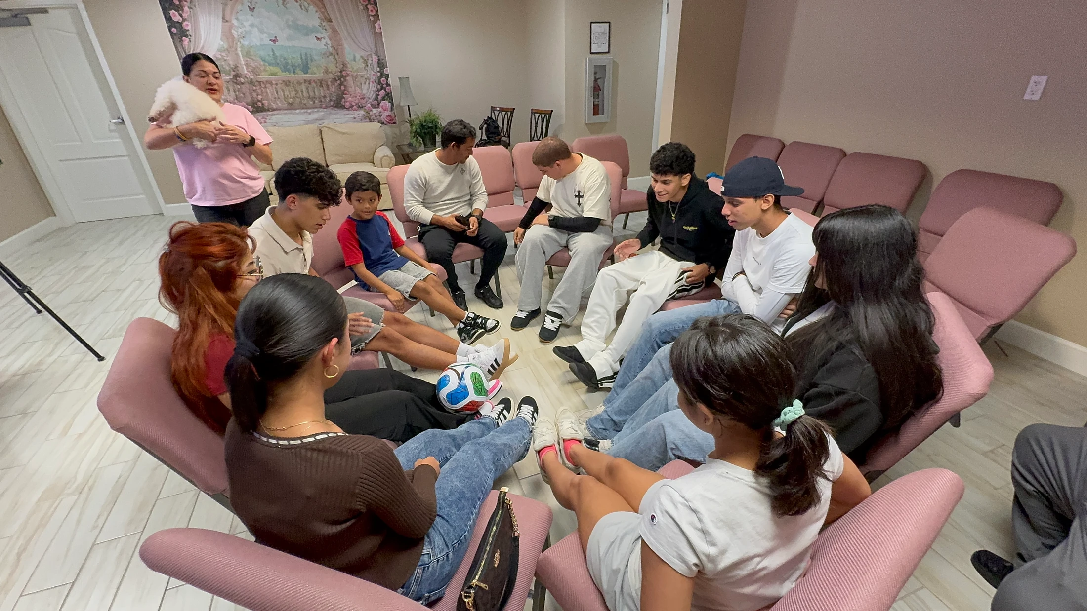 A diverse group of young people are sitting in a circle of pink chairs indoors, engaged in a conversation, with a soccer ball on the floor in the middle.