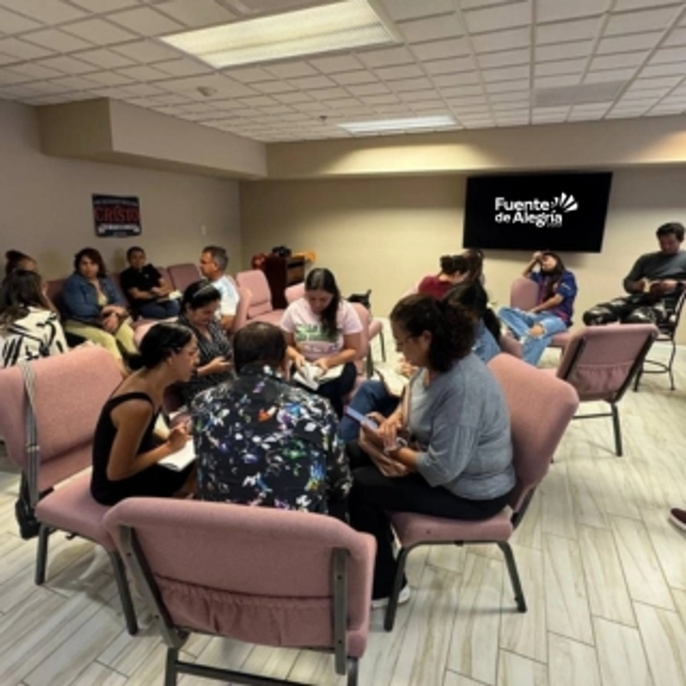 A group of adults sits in chairs arranged in a circle inside a meeting room, engaged in conversation. A screen in the background shows a logo.