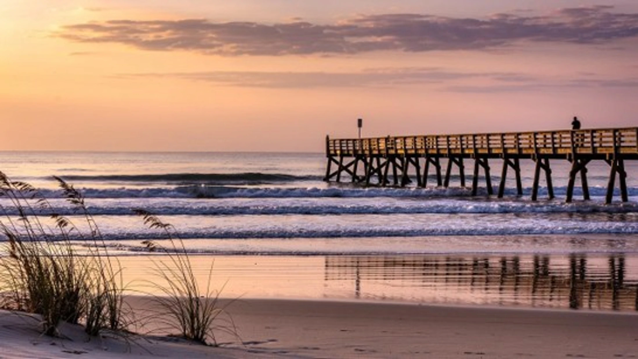 A wooden pier stretches into the ocean under a soft pink and orange sunrise sky. A lone person stands at the far end as gentle waves roll onto the sandy beach.