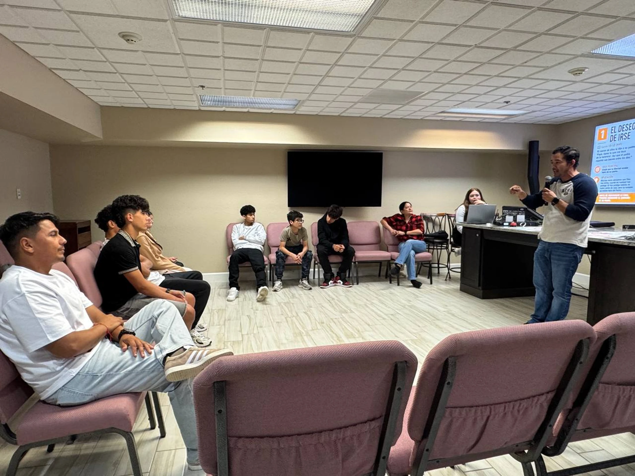 A man in a baseball shirt and jeans speaks into a microphone, presenting to a group of young people seated in pink chairs in a meeting room.