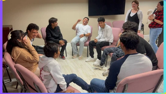 A group of teenagers sit in a circle on pink chairs, laughing and talking together during a meeting. Two women stand nearby, one holding a small white dog.