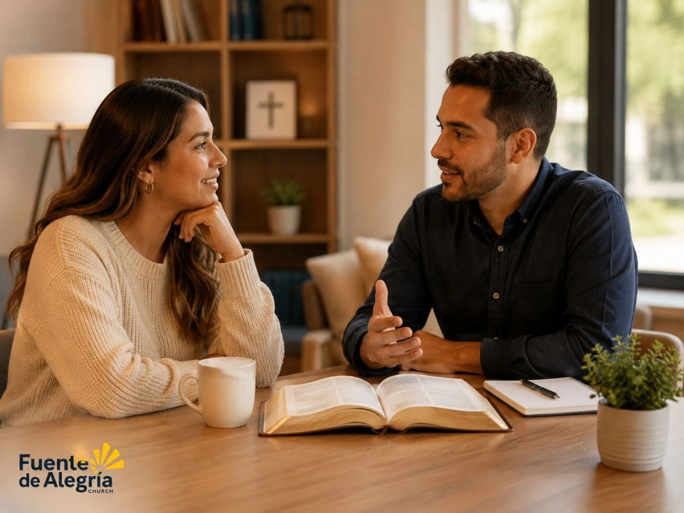 A man and a woman sit at a wooden table with an open Bible between them, looking at each other and smiling as they talk in a cozy living room.