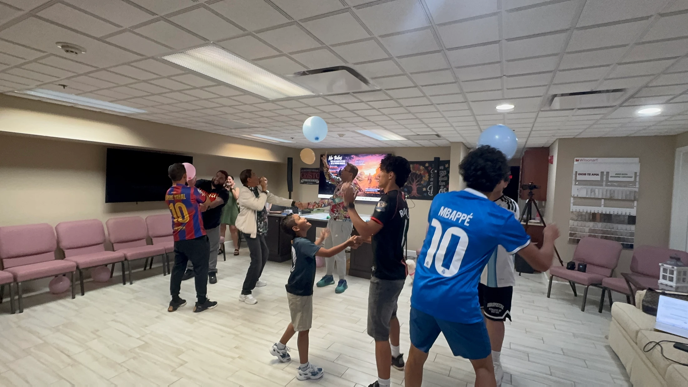 A diverse group of adults and children laugh and play together, trying to keep blue and pink balloons in the air inside a large community room.