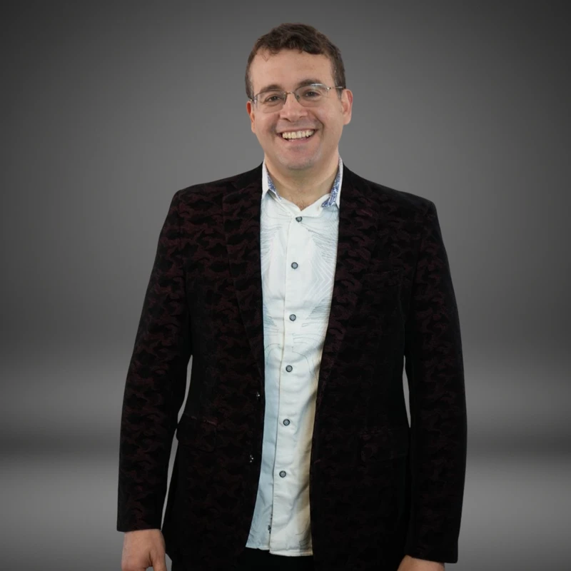 A waist-up studio portrait of a smiling man with glasses, wearing a dark patterned blazer over a light-colored button-up shirt against a gray background.