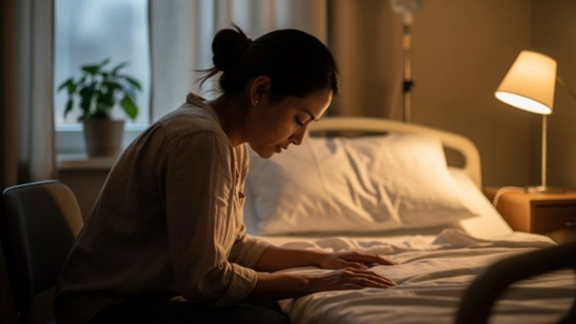 A sad woman with her hair in a bun sits in a dimly lit hospital room, leaning over an empty hospital bed with her head bowed and hands resting on the sheets.