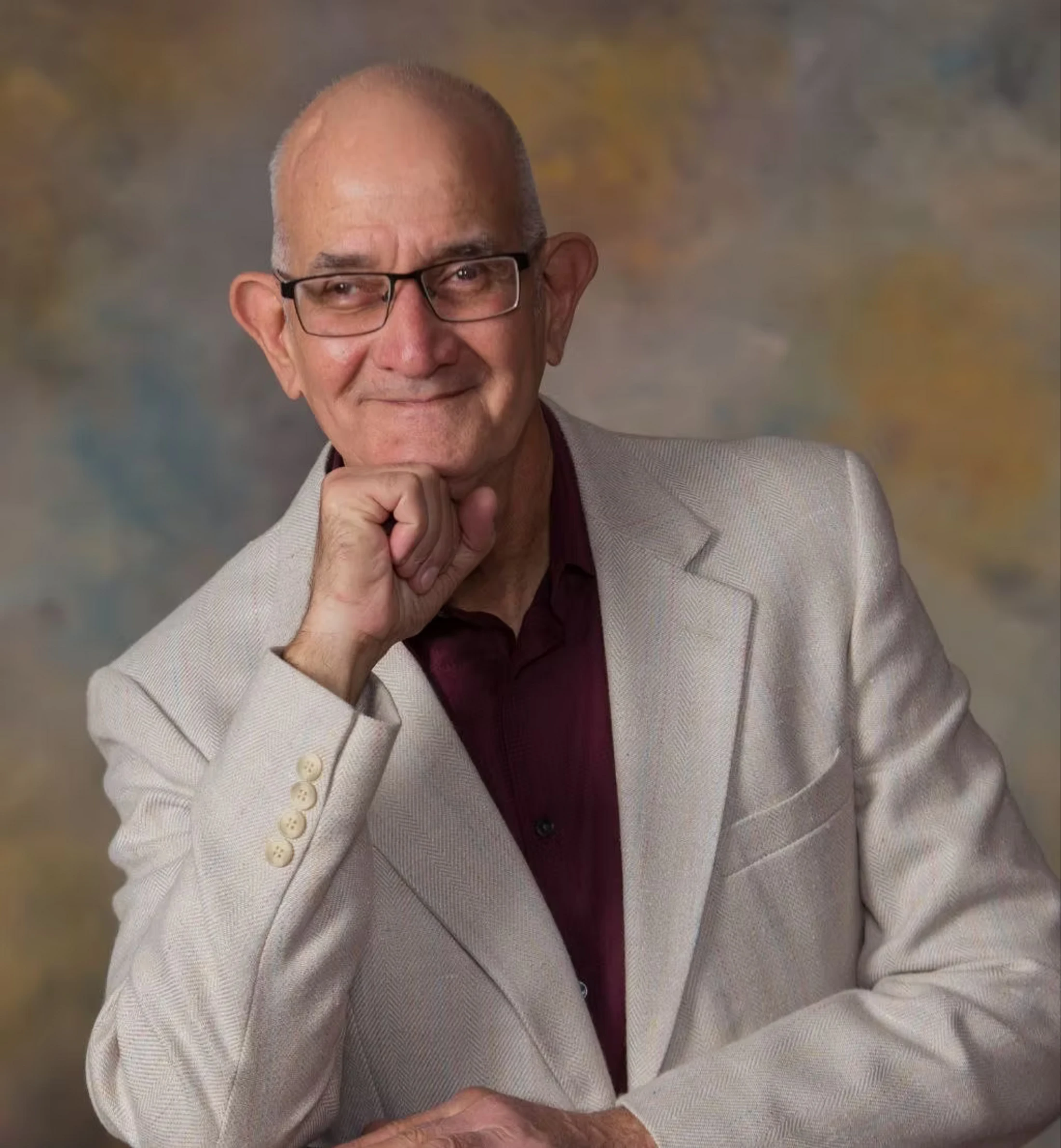 A professional headshot of an older, bald man with glasses, smiling gently. He wears a light-colored blazer over a dark shirt and rests his chin on his fist.