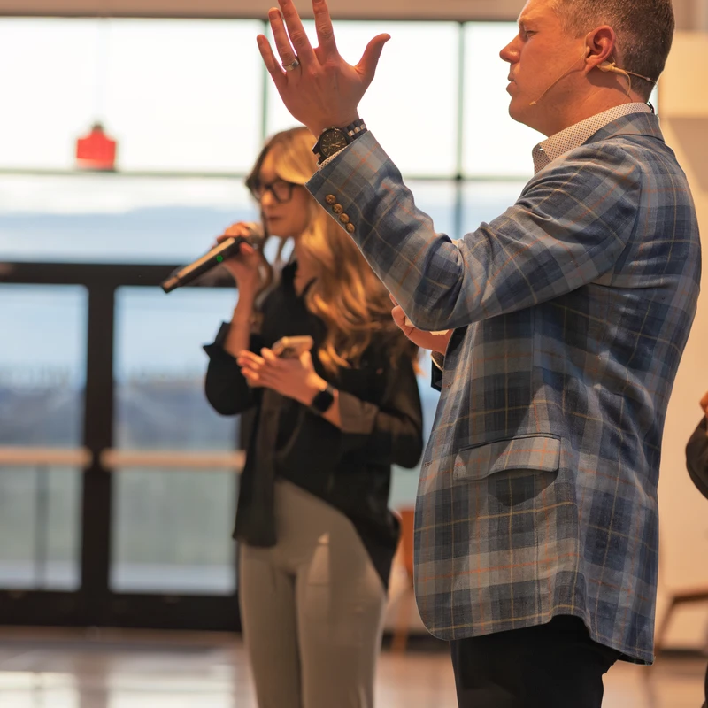 A man in a blue plaid blazer raises his hand while speaking. He wears a headset microphone. A woman with a microphone stands blurred in the background.