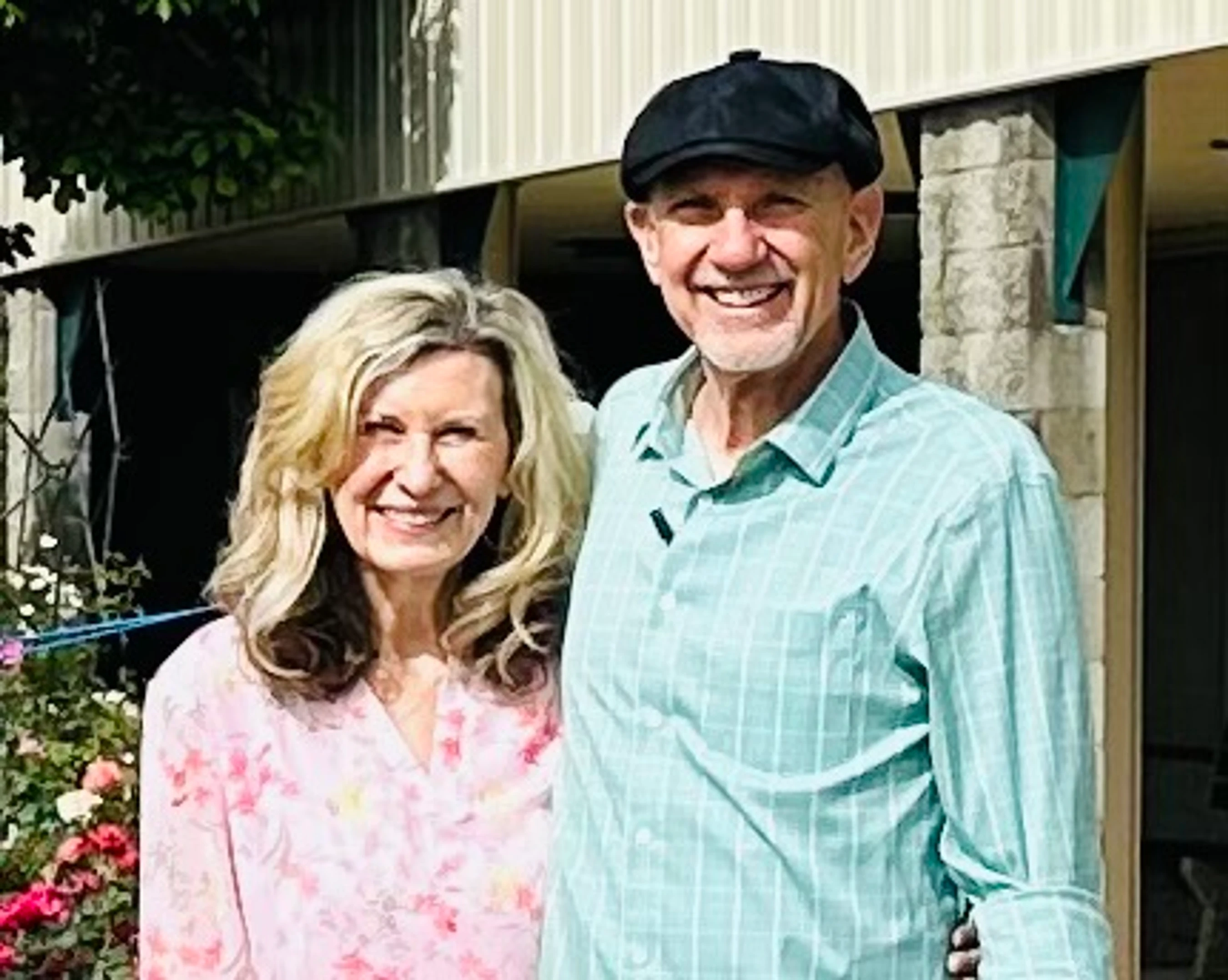 A man and woman stand close together, smiling at the camera. He wears a light blue plaid shirt and a black cap; she wears a pink floral blouse.