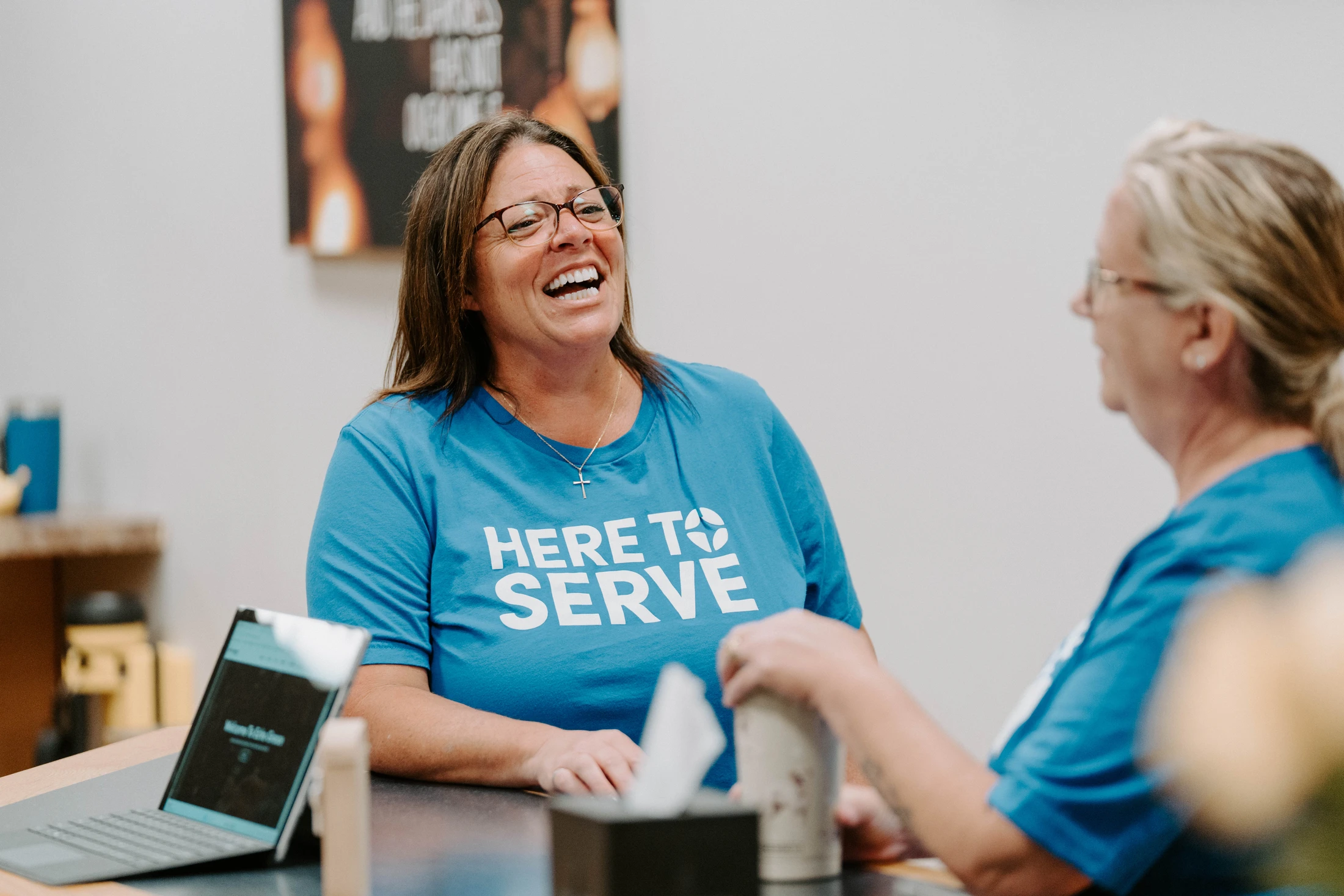 A woman with glasses wearing a blue t-shirt that says 'Here to Serve' is laughing while sitting at a desk talking with another person.