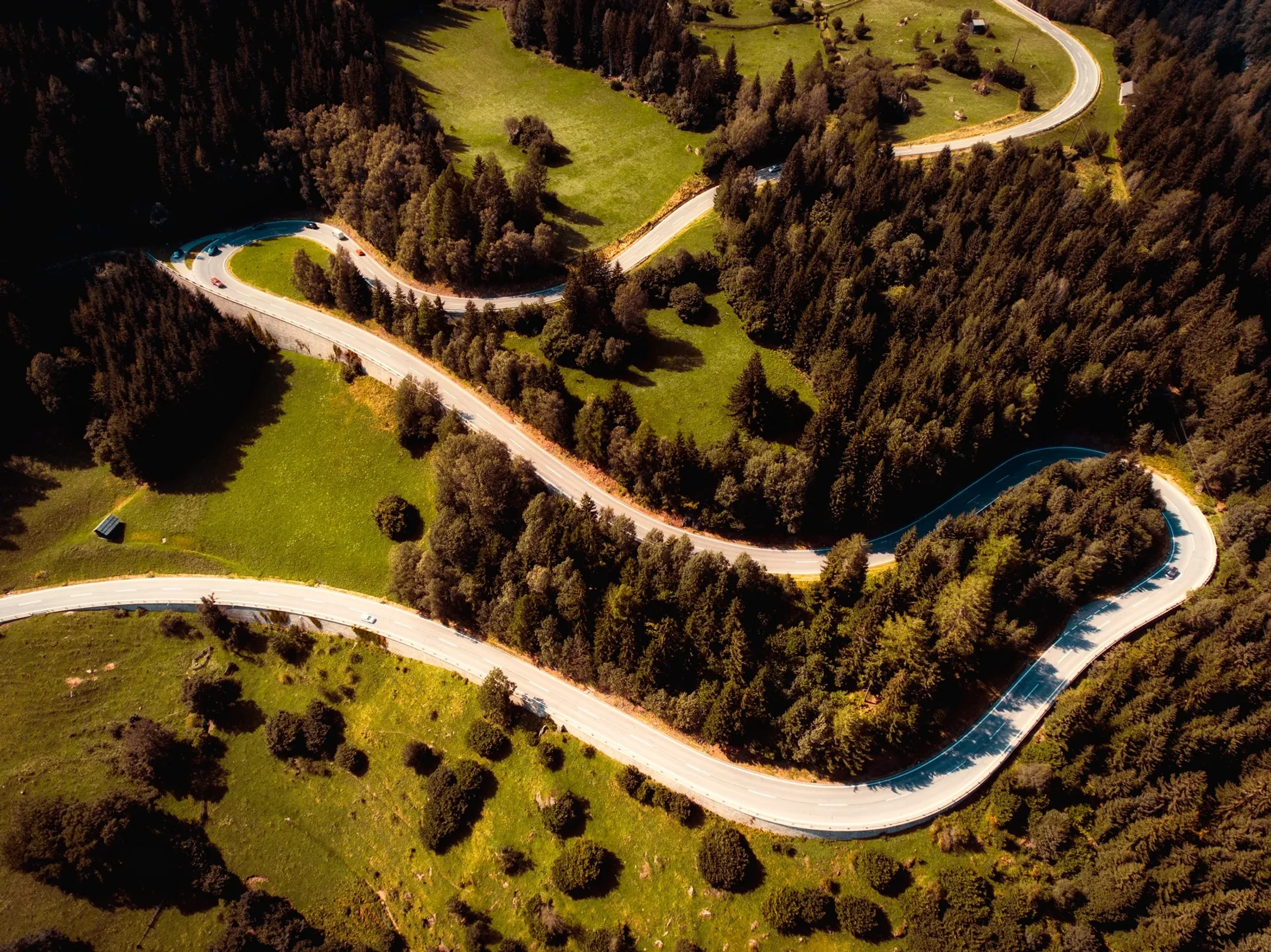 An aerial, high-angle view of a winding road with sharp turns snaking through a dense forest and green, grassy fields. Several cars drive on the road.