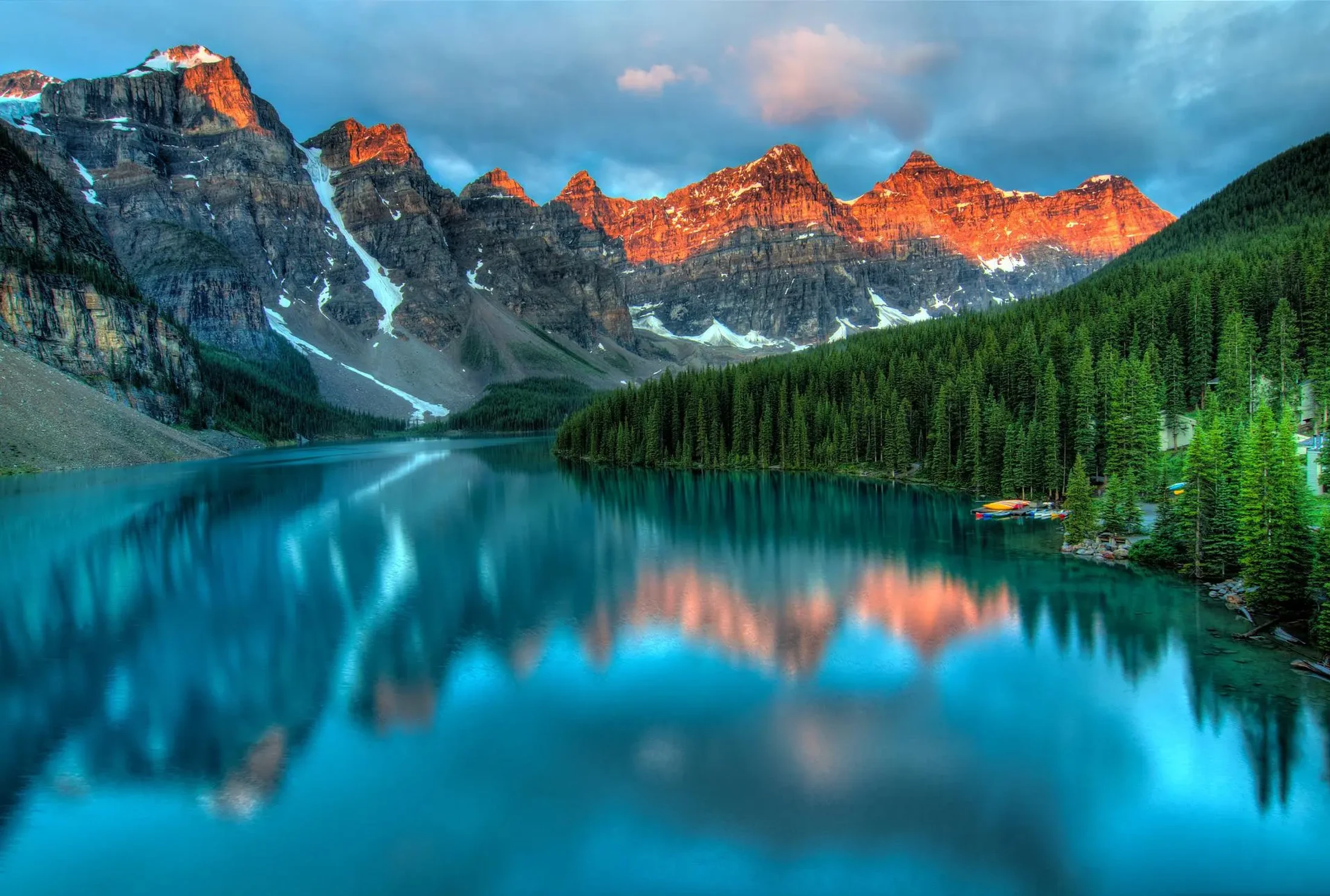 A serene turquoise lake reflects a majestic mountain range with snow-dusted peaks glowing orange from the sunrise, bordered by a dense evergreen forest.