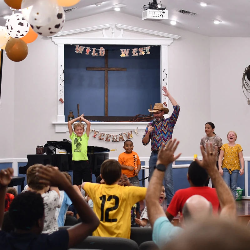 Children and adults on stage at a Wild West themed church event, leading an enthusiastic audience of kids who are cheering and raising their hands.