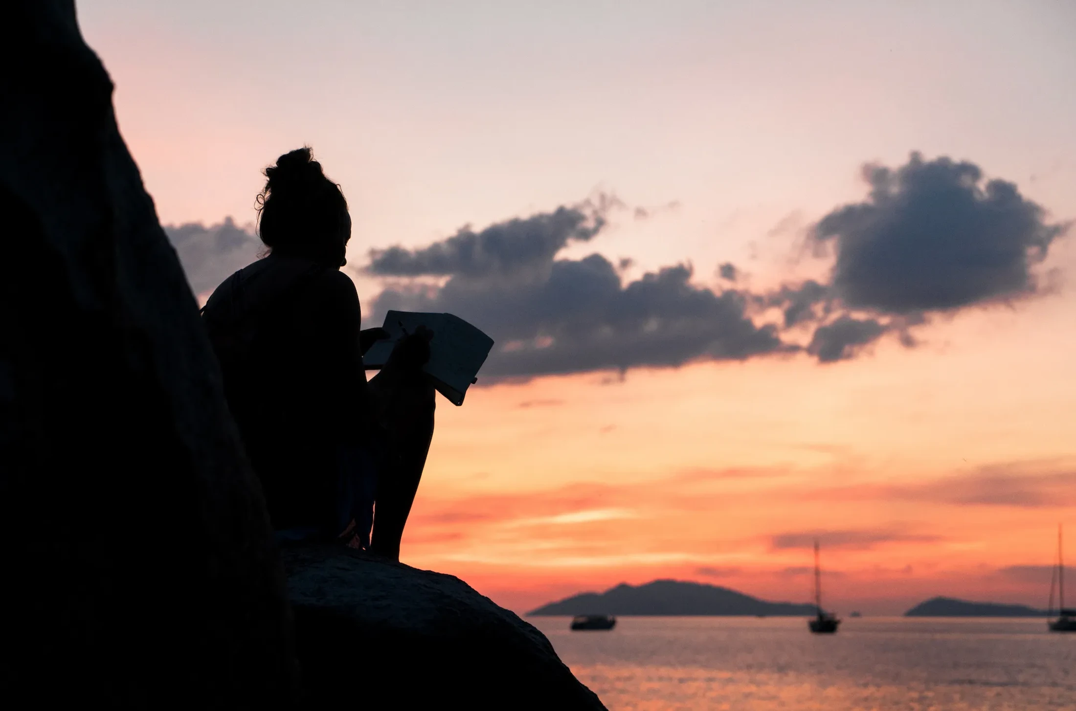 A silhouette of a person sitting on a rock and reading a book against a beautiful sunset sky over the sea, with distant boats and islands.