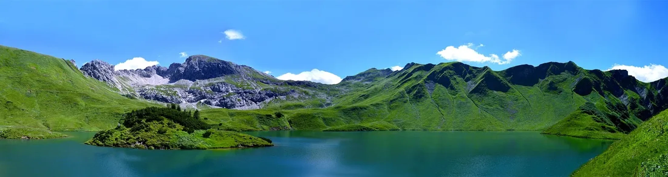 A wide panoramic photo of a calm, blue-green alpine lake surrounded by rolling, bright green mountains under a clear blue sky with a few white clouds.