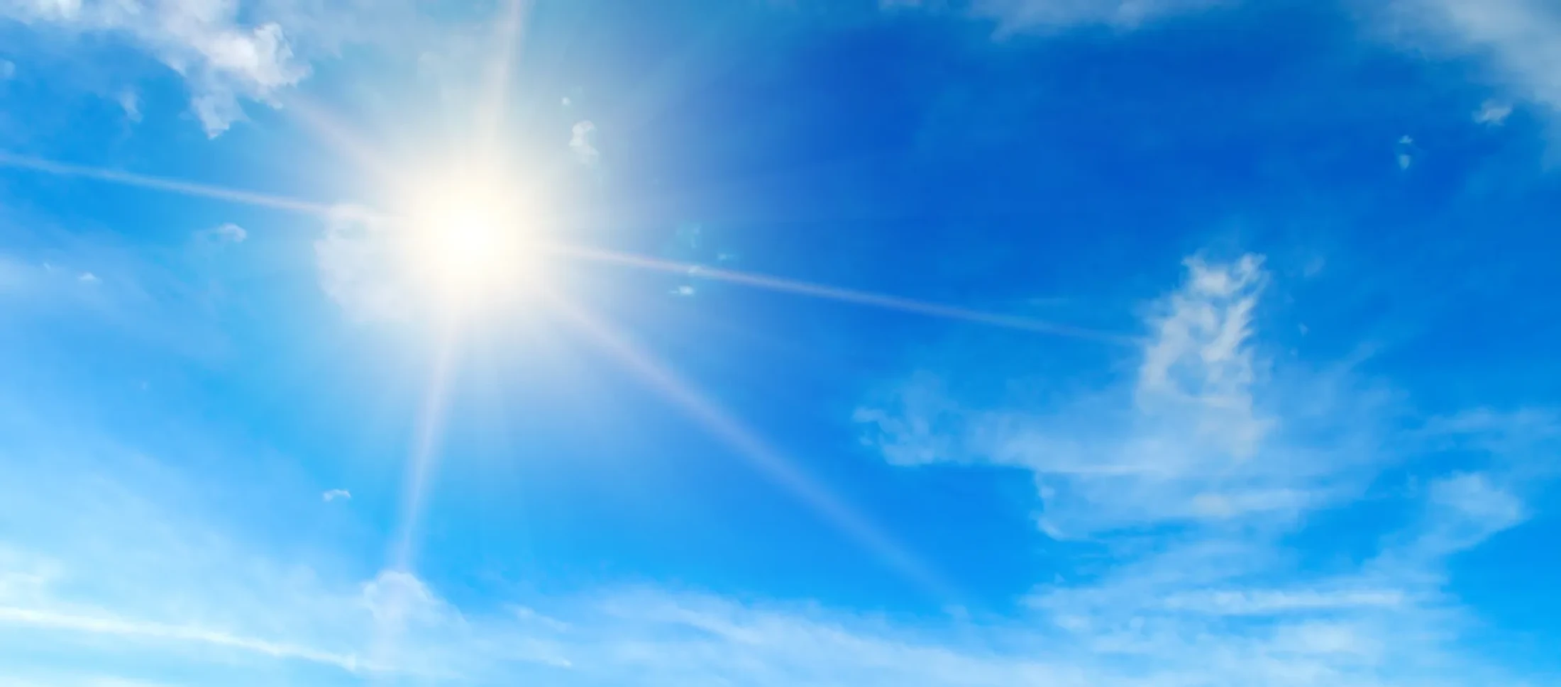 The bright sun shines in a vibrant blue sky, with rays of light spreading out over a few scattered, wispy white clouds.