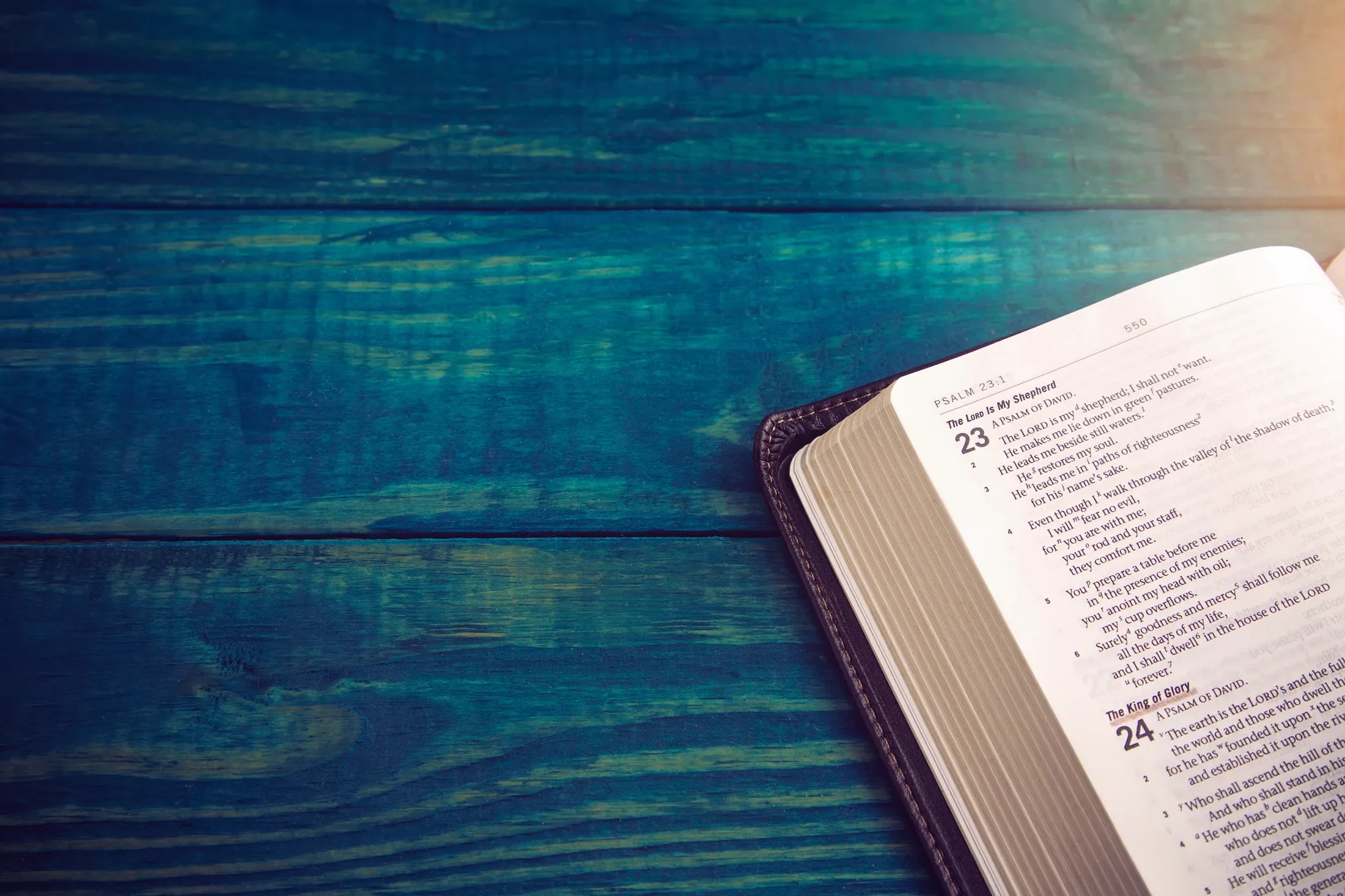 An open Bible with a dark leather cover rests on a teal wooden surface. The page is open to Psalm 23, with warm light highlighting the text.