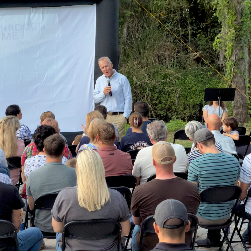 A man addresses a seated audience during an outdoor event. He holds a microphone and stands before a large white screen with Christian lyrics.