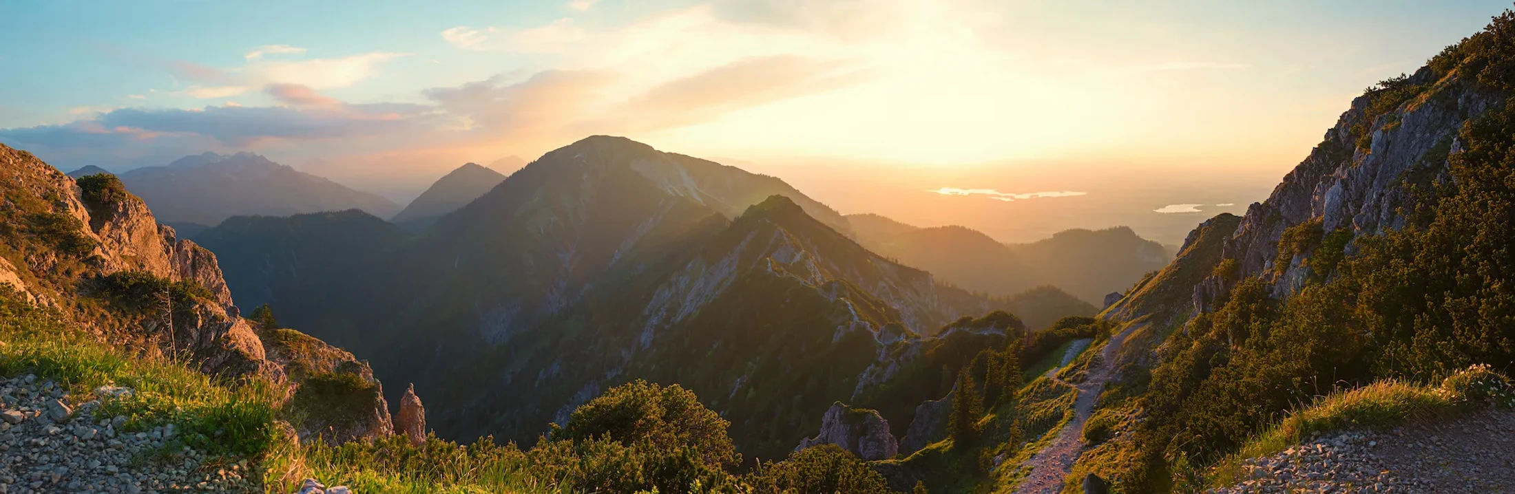 A sweeping panoramic view of a mountain range bathed in the warm, golden light of sunrise, with a hiking path winding along a ridge in the foreground.
