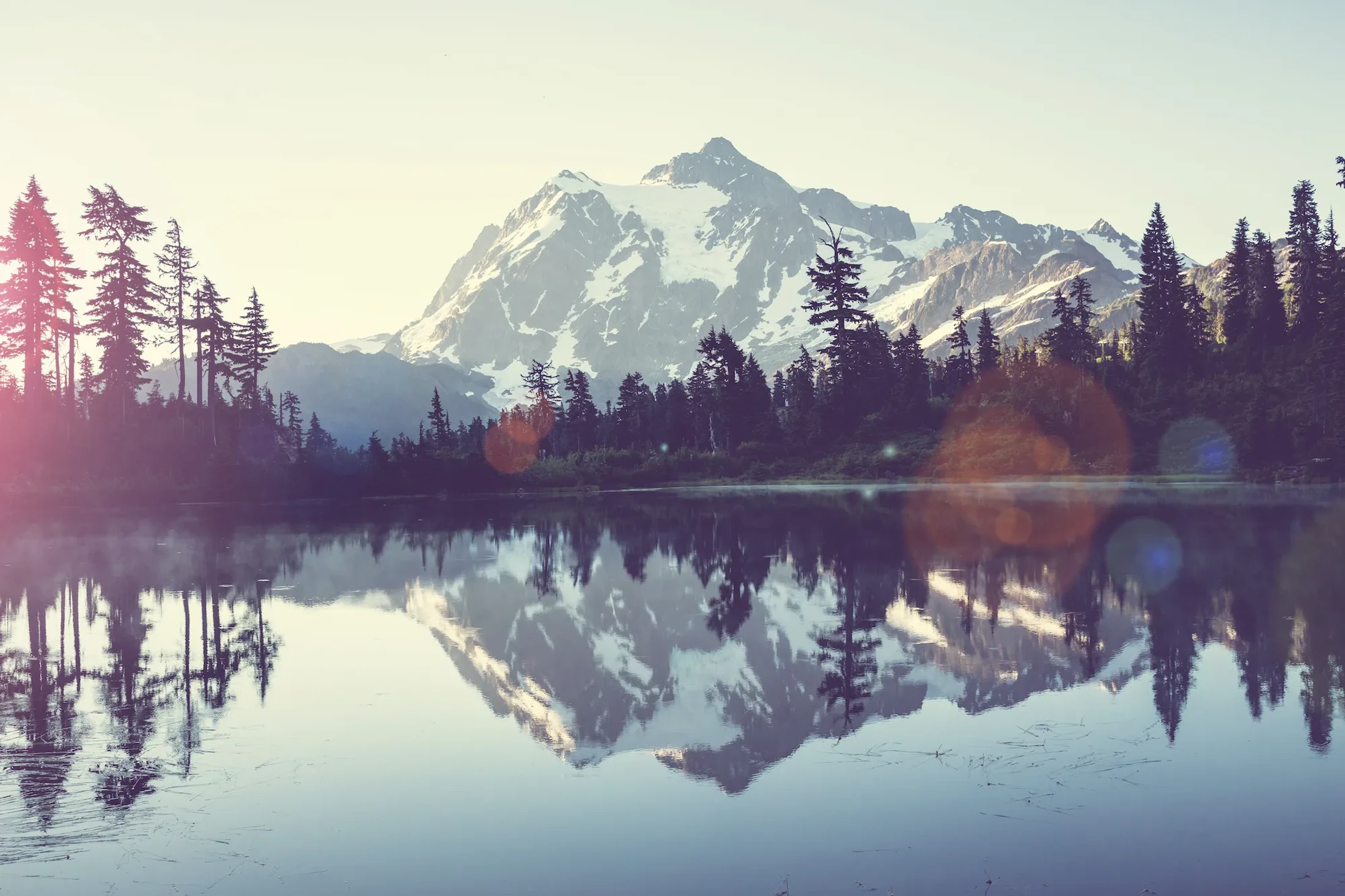 A snow-capped mountain and a pine forest are perfectly reflected in the still water of a lake, creating a symmetrical view under a hazy sky with lens flare.