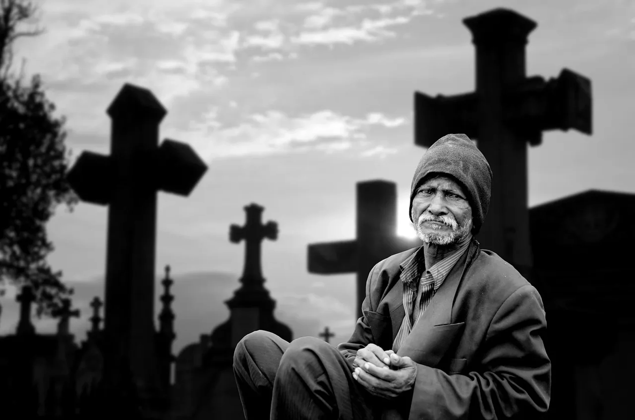 A black and white photo of an elderly man wearing a beanie and jacket, sitting in a cemetery with large, silhouetted crosses in the background.