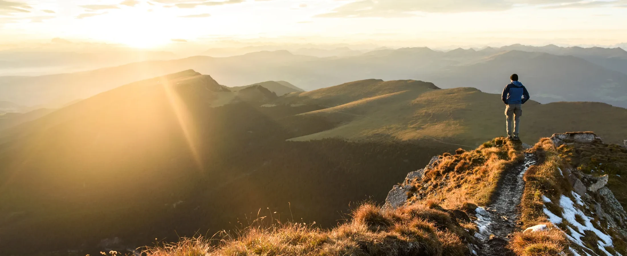 A hiker stands on a mountain summit, looking out at a panoramic view of rolling mountains bathed in the warm, golden light of a sunrise or sunset.