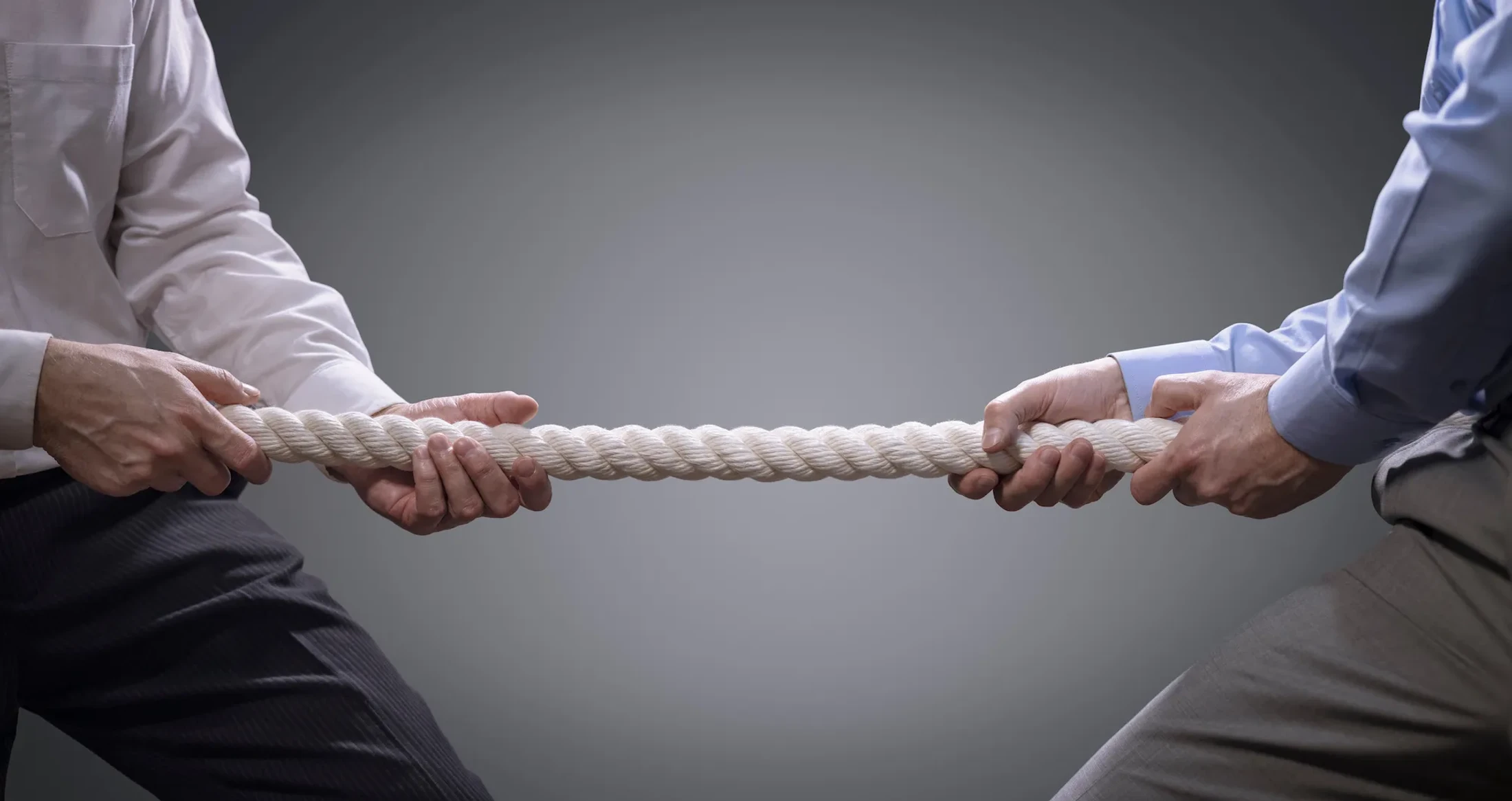Two men in dress shirts and slacks pull on opposite ends of a thick rope in a game of tug of war, representing business competition against a gray background.