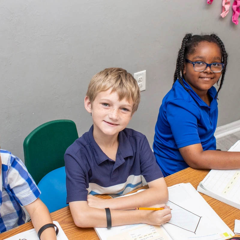 Three diverse children sit at a desk doing their schoolwork. A boy and a girl look up from their books and smile at the camera.
