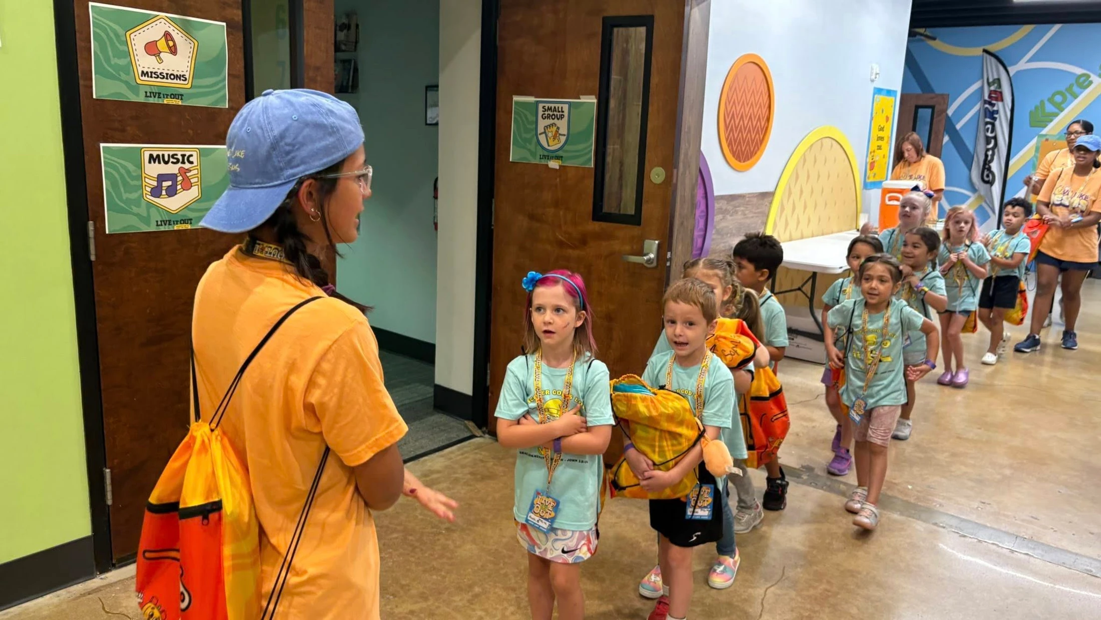 A female camp counselor in an orange shirt talks to a line of young children wearing matching blue shirts in a brightly decorated hallway.
