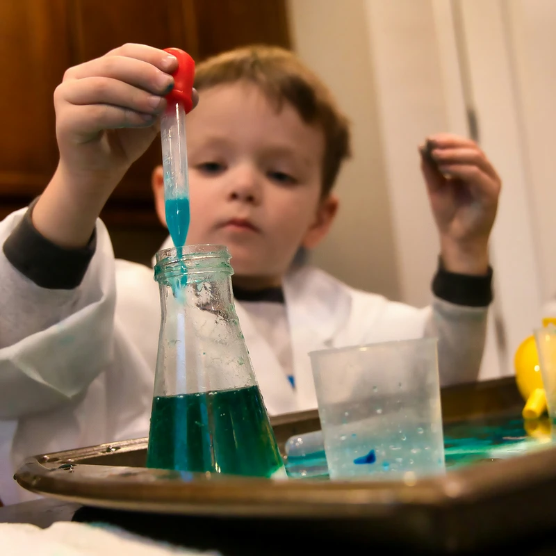 A young boy in a white lab coat focuses intently as he uses a pipette to transfer blue liquid into a glass flask as part of a science experiment.