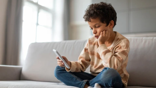 A young boy with curly hair sits cross-legged on a grey sofa, looking at a smartphone. He wears a striped sweater and rests his chin on his hand.