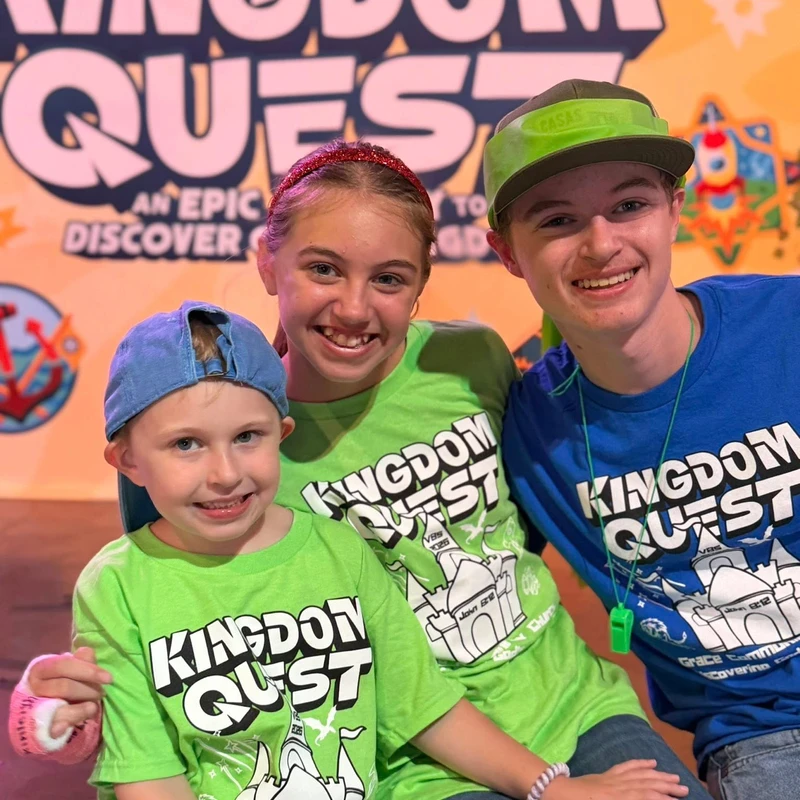 Three smiling children, a girl between two boys, wear matching 'Kingdom Quest' t-shirts in green and blue while posing in front of an event banner.