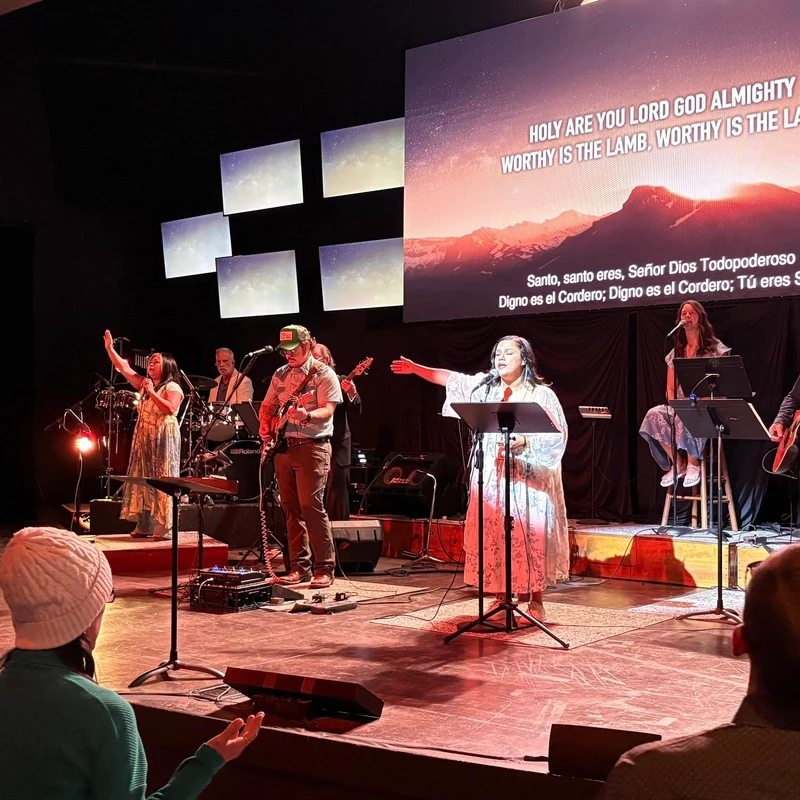 A diverse band performs on a warmly lit stage, leading a song. A large screen behind them displays lyrics in English and Spanish over a mountain scene.