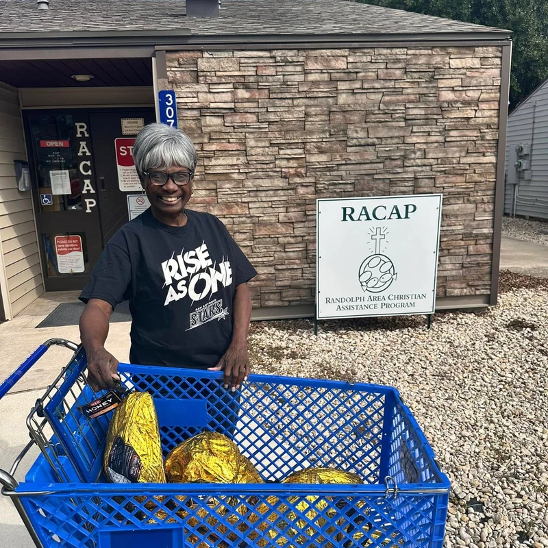 A smiling woman stands outside a building with a blue shopping cart filled with hams, donating to the Randolph Area Christian Assistance Program.
