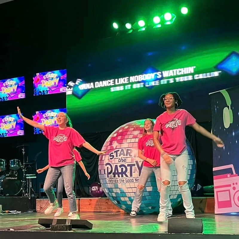A group of young women in matching pink t-shirts and jeans dance energetically on a stage with a large disco ball and screens that say 'Start the Party'.