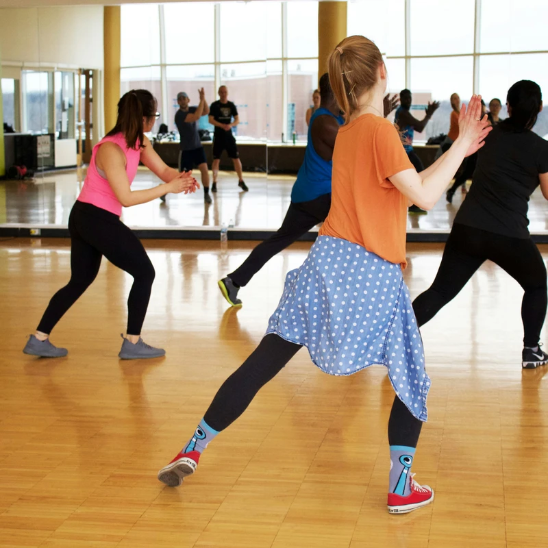 A group of diverse people in workout attire participate in a dance fitness class inside a bright studio with a hardwood floor and a wall of mirrors.