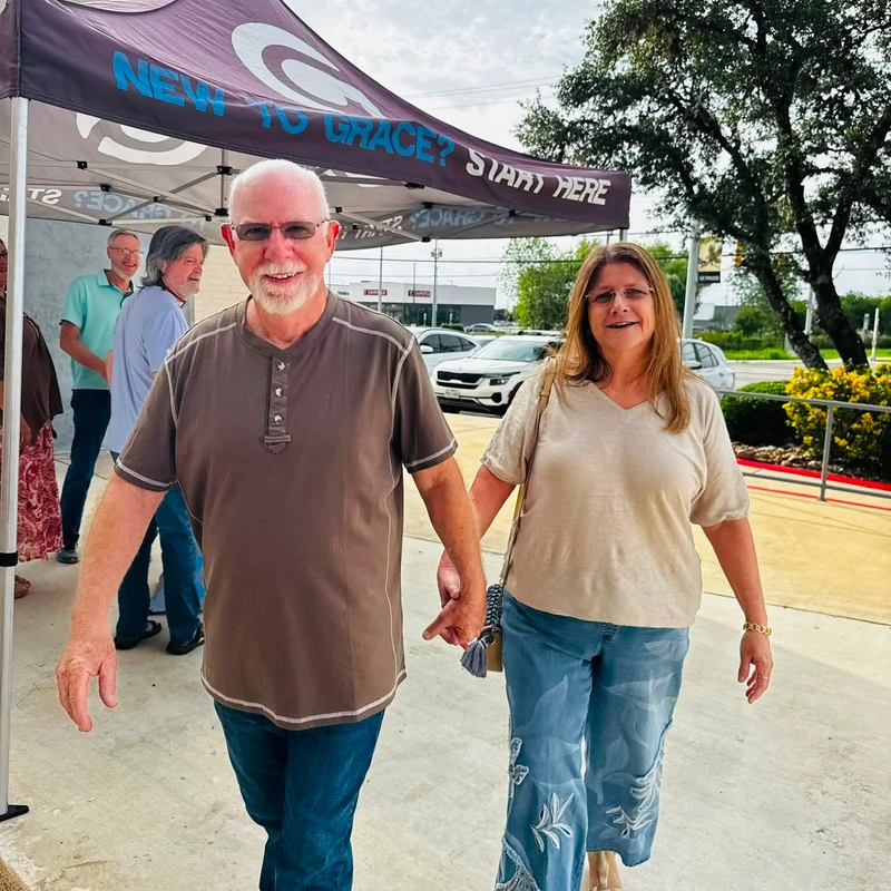 An older man and woman smile and hold hands as they walk toward the camera. In the background, there is a purple welcome tent that says 'New to Grace?'.