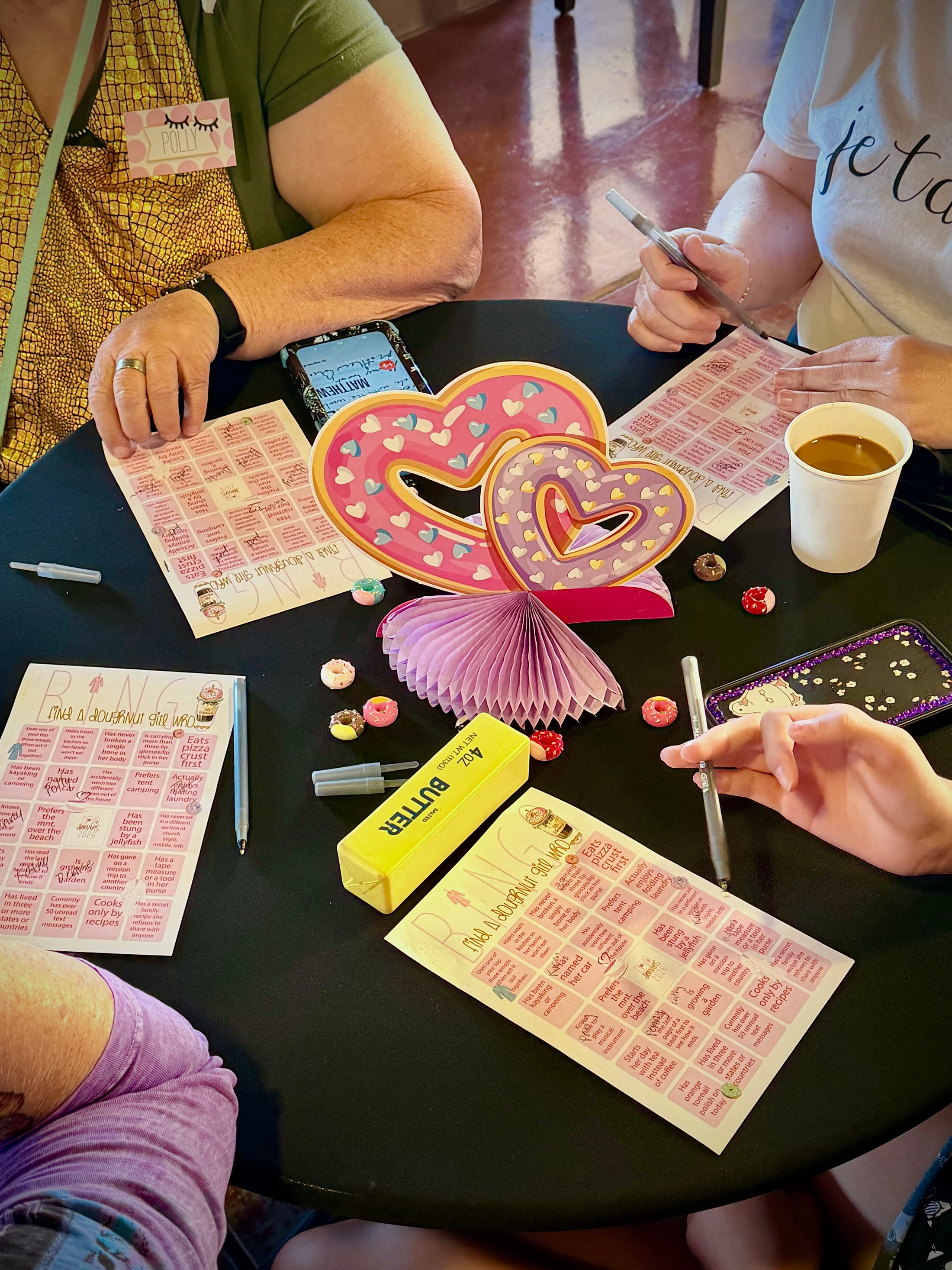 Three people sit around a black table playing a doughnut-themed bingo game. The table is decorated with a heart centerpiece, coffee, and small doughnut charms.