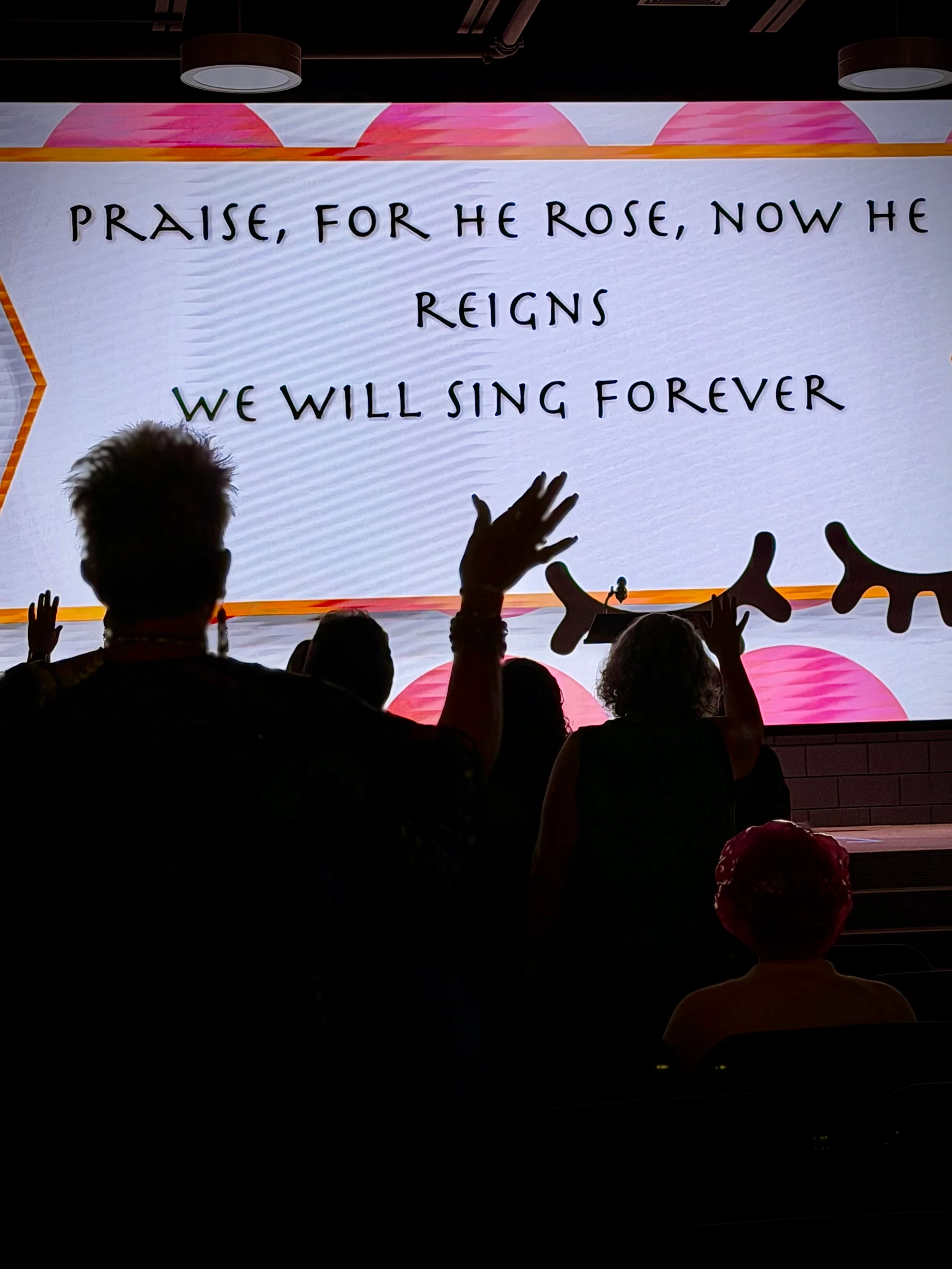 Silhouettes of a congregation with hands raised in worship, facing a screen with the lyrics 'PRAISE, FOR HE ROSE, NOW HE REIGNS WE WILL SING FOREVER'.