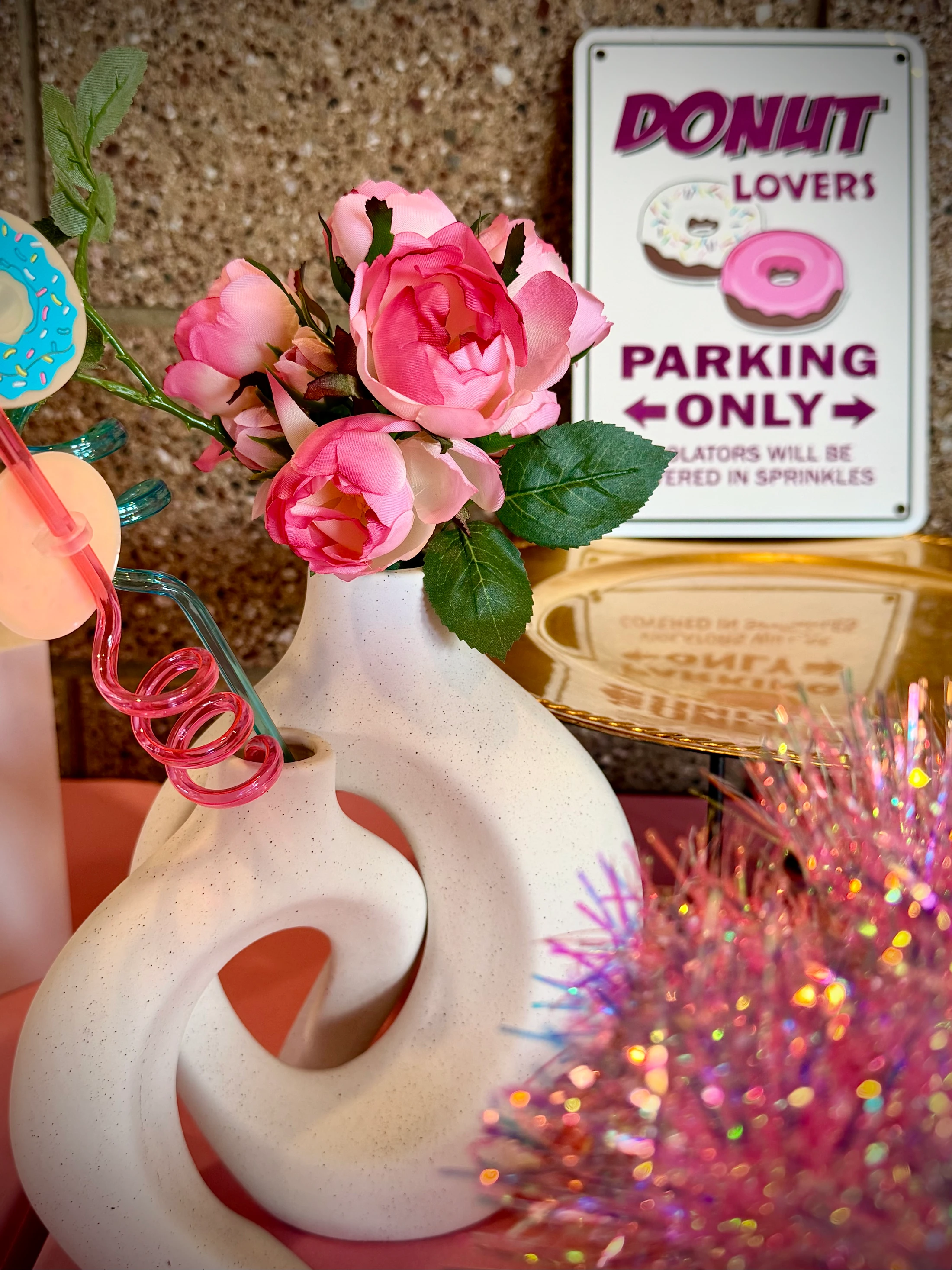 A white abstract vase holds pink artificial flowers and colorful straws, next to a novelty sign that reads 'Donut Lovers Parking Only'.