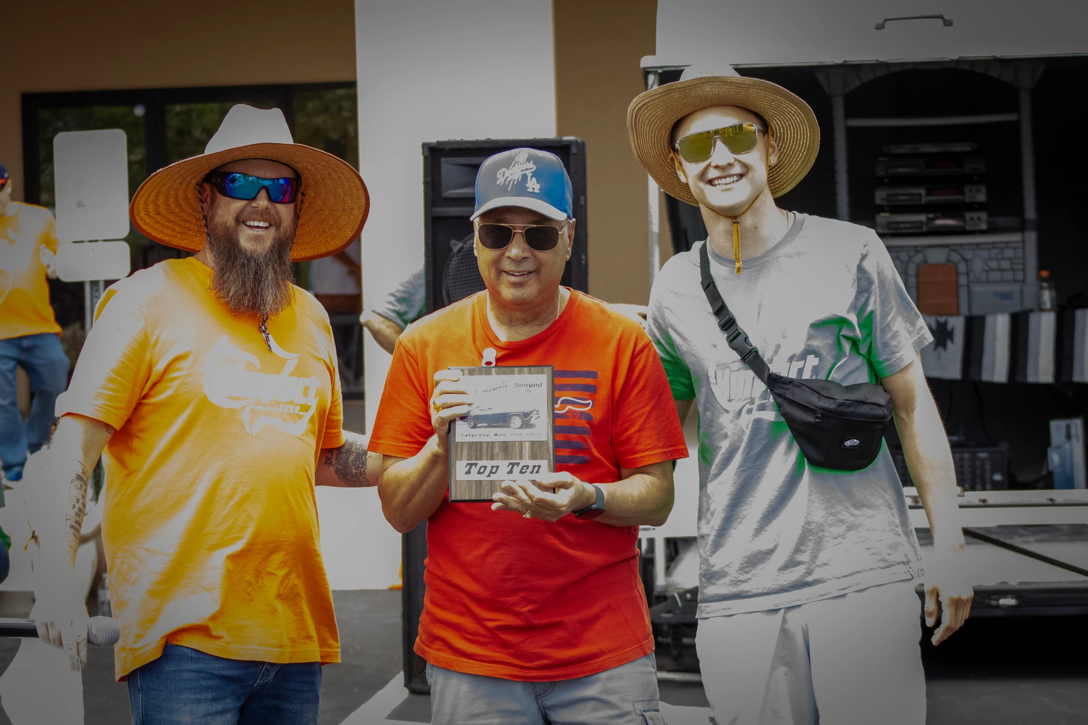 Three men smile for a photo outdoors. The man in the middle holds a 'Top Ten' plaque, while the men on either side wear sunglasses and straw hats.