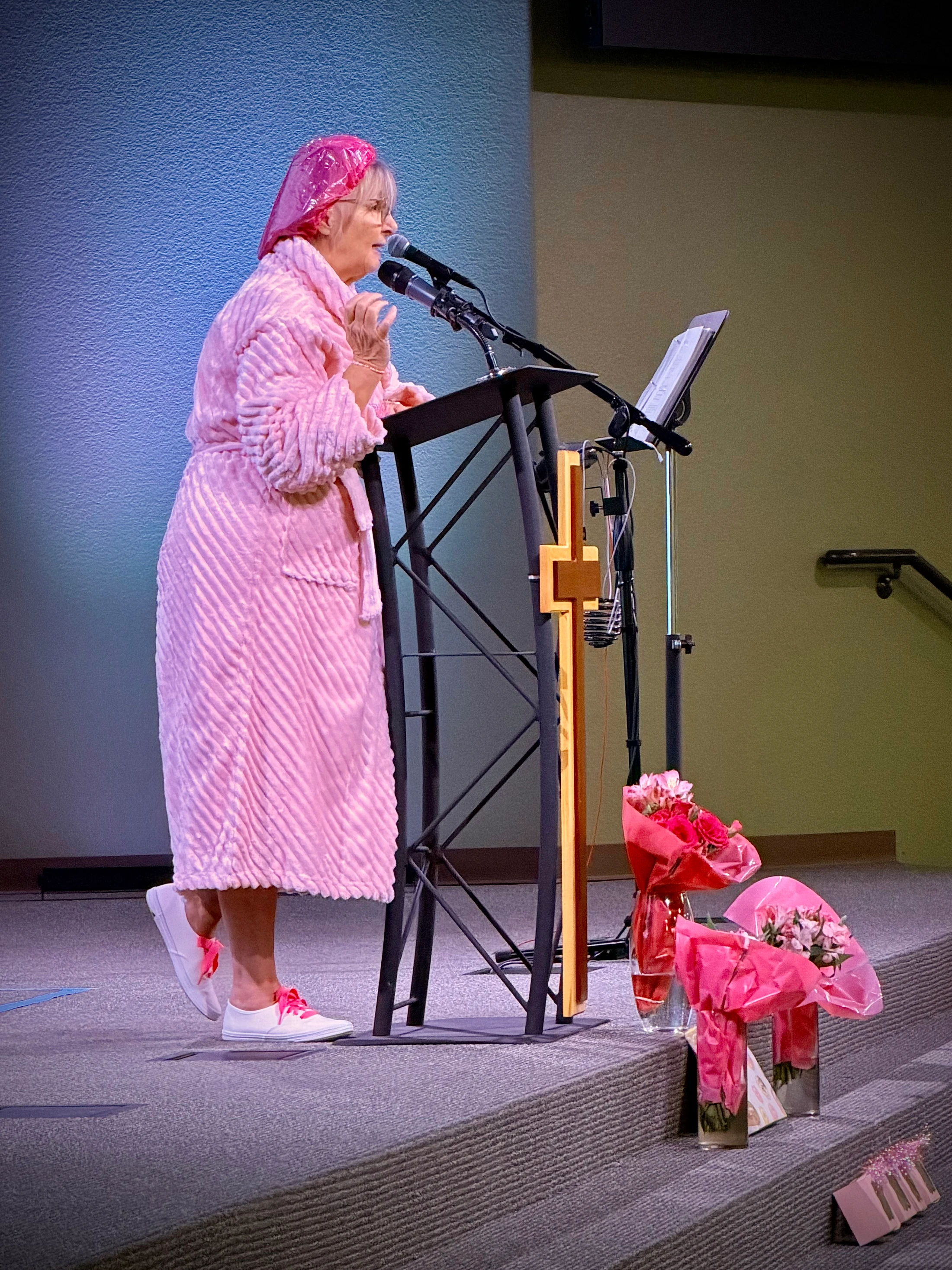 A woman wearing a pink bathrobe, shower cap, and sneakers with pink laces speaks at a lectern on a stage. Bouquets of flowers are on the steps below.
