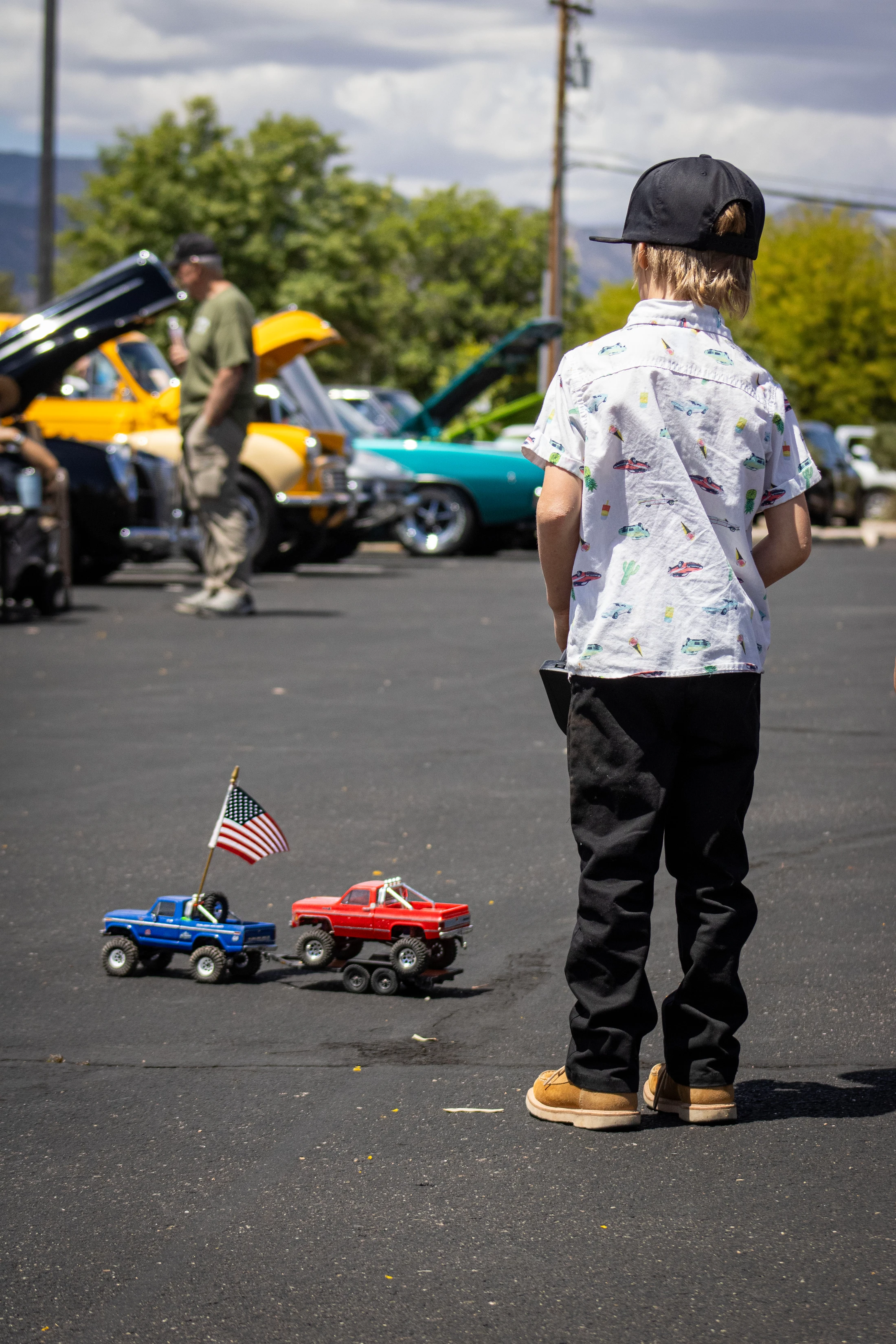 A boy, seen from behind, stands at a car show with his toy monster trucks. A blue truck with an American flag tows a red truck on a trailer.