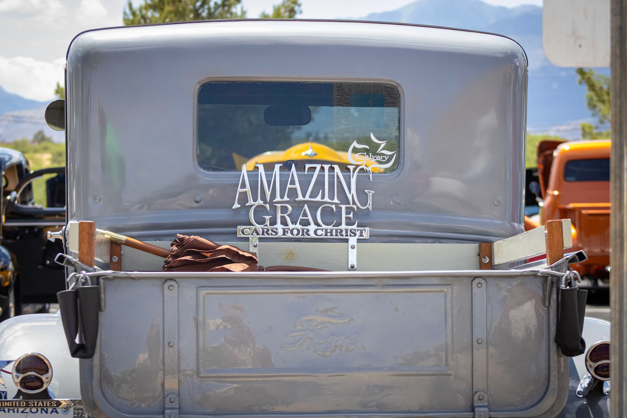The back of a vintage gray Ford pickup truck with a chrome emblem that reads 'Amazing Grace, Cars for Christ'. A brown umbrella rests in the truck bed.