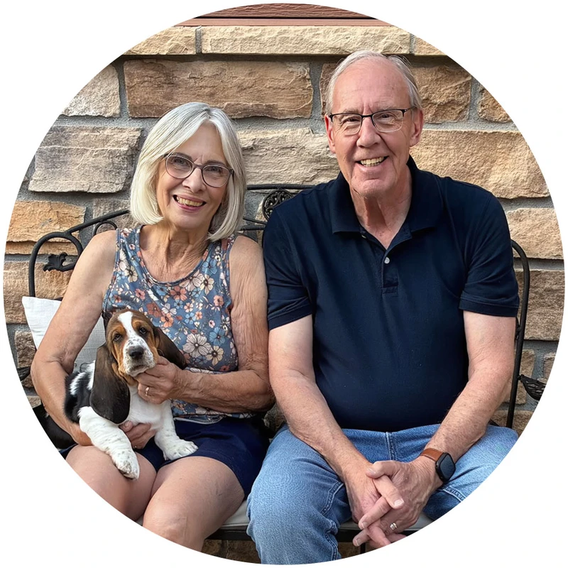 A smiling older man and woman sit on a bench in front of a stone wall. The woman is holding a small brown, black, and white Basset Hound puppy on her lap.