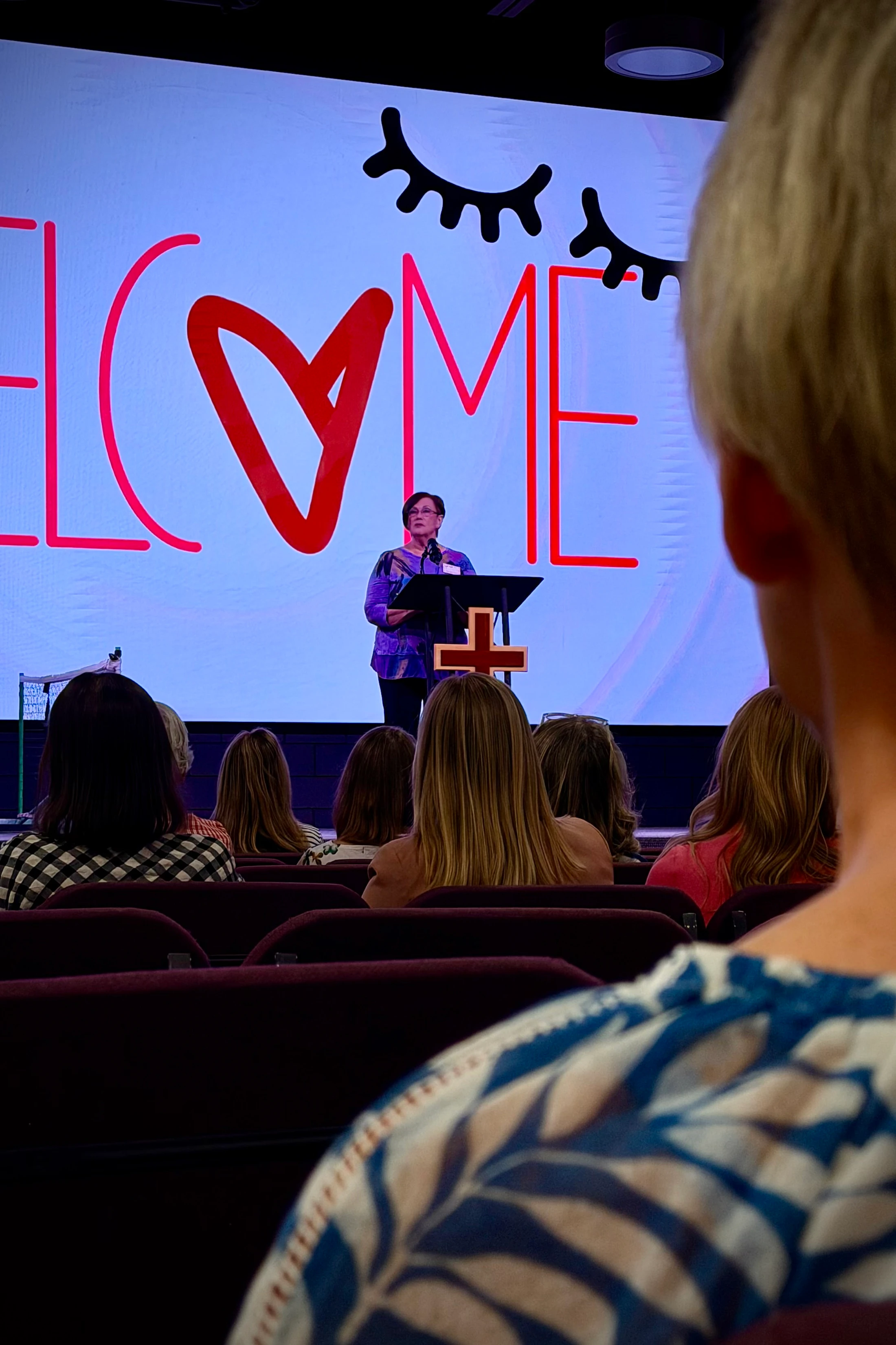 An over-the-shoulder view of an audience listening to a woman speak at a church podium. A large screen behind her says 'WELCOME' with a heart for the letter O.